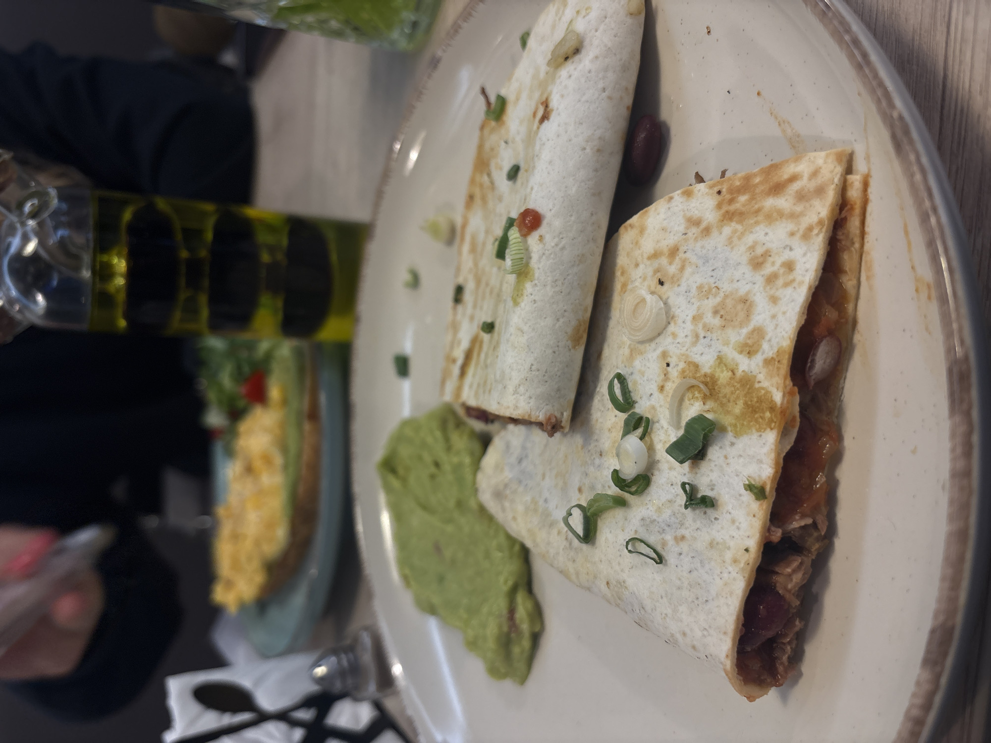 A plate of quesadillas sits on a wooden table. The quesadillas are cut into triangular halves and appear to be filled with a mixture of meat and beans, topped with green onions. A dollop of guacamole is beside the quesadillas. In the background, there's a salad in a glass bowl, a bottle of oil, a person holding a phone, and a set of cutlery on the table. The location appears to be a restaurant in Varna, Bulgaria.