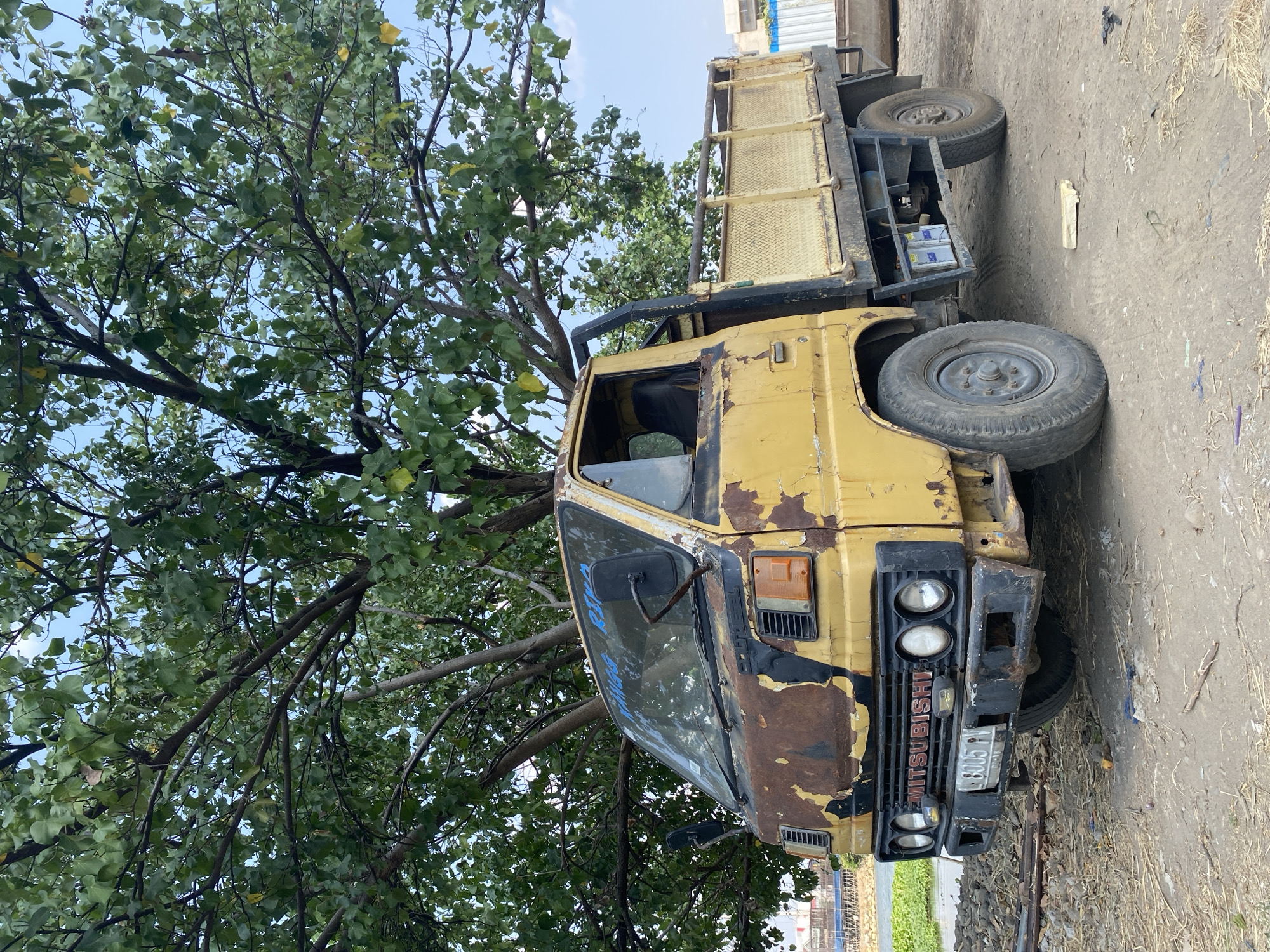 A yellow Mitsubishi truck, heavily rusted and weathered, is parked on a dirt lot under the shade of a large, leafy tree. The truck has a flatbed with yellow railings and two visible batteries mounted underneath. The front of the truck shows significant rust and peeling paint, with round headlights and a prominent "MITSUBISHI" logo on the grille. A license plate reading "B 8005 PV" is affixed to the front bumper. In the background, a body of water and buildings can be seen, suggesting an urban or semi-urban environment, possibly near the river in Kediri City, Indonesia. The sky is partly cloudy.