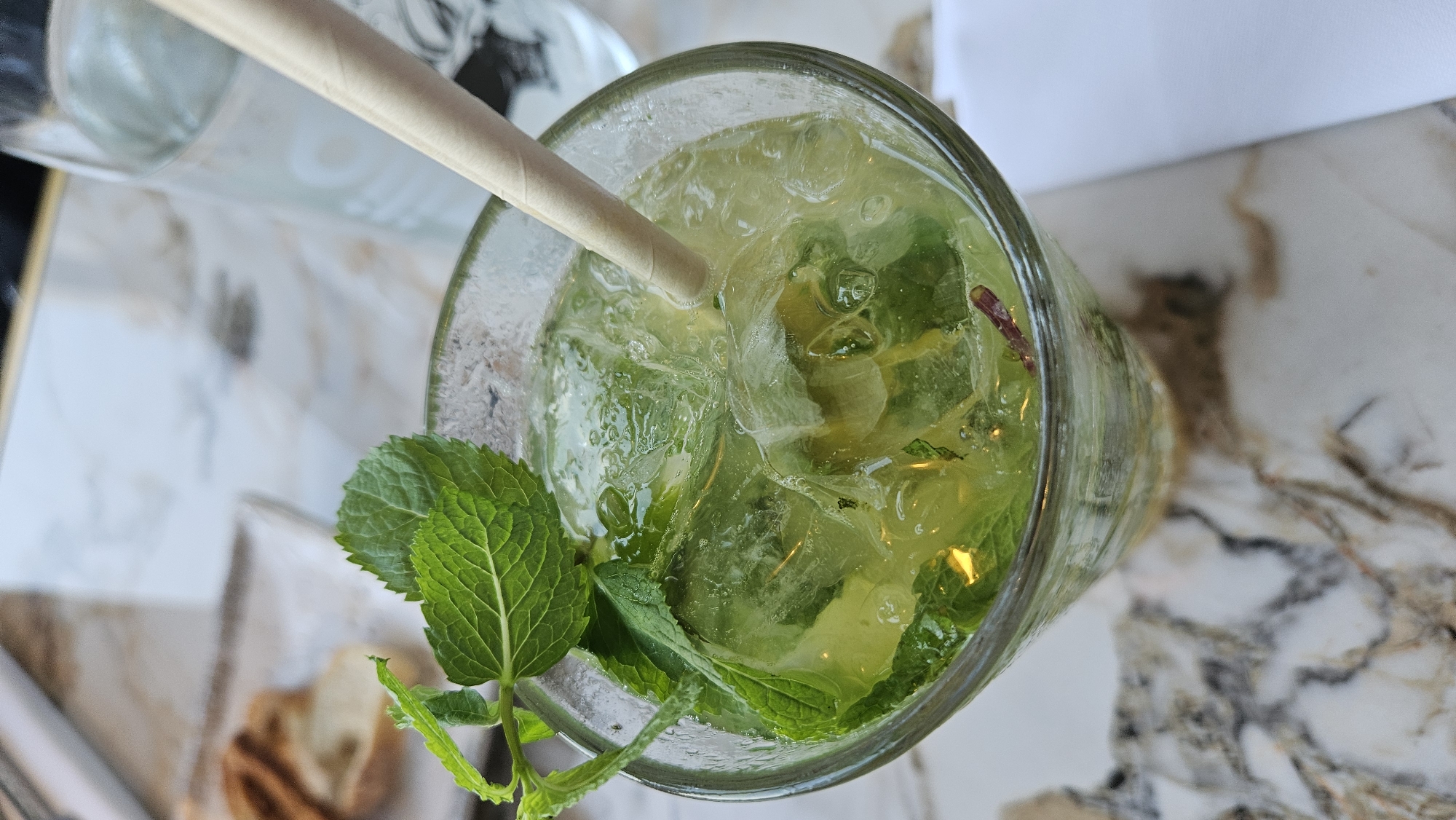 A close-up, top-down shot focuses on a refreshing beverage in a clear glass filled with ice, mint, and what appears to be lime. A light-colored paper straw extends from the drink. The glass rests on a marbled surface. In the background, slightly out of focus, is a glass of water and a plate with some toasted bread. The scene is likely set in L'Île-Rousse, France, possibly at a cafe or restaurant given the presentation. There are no people visible in the frame.