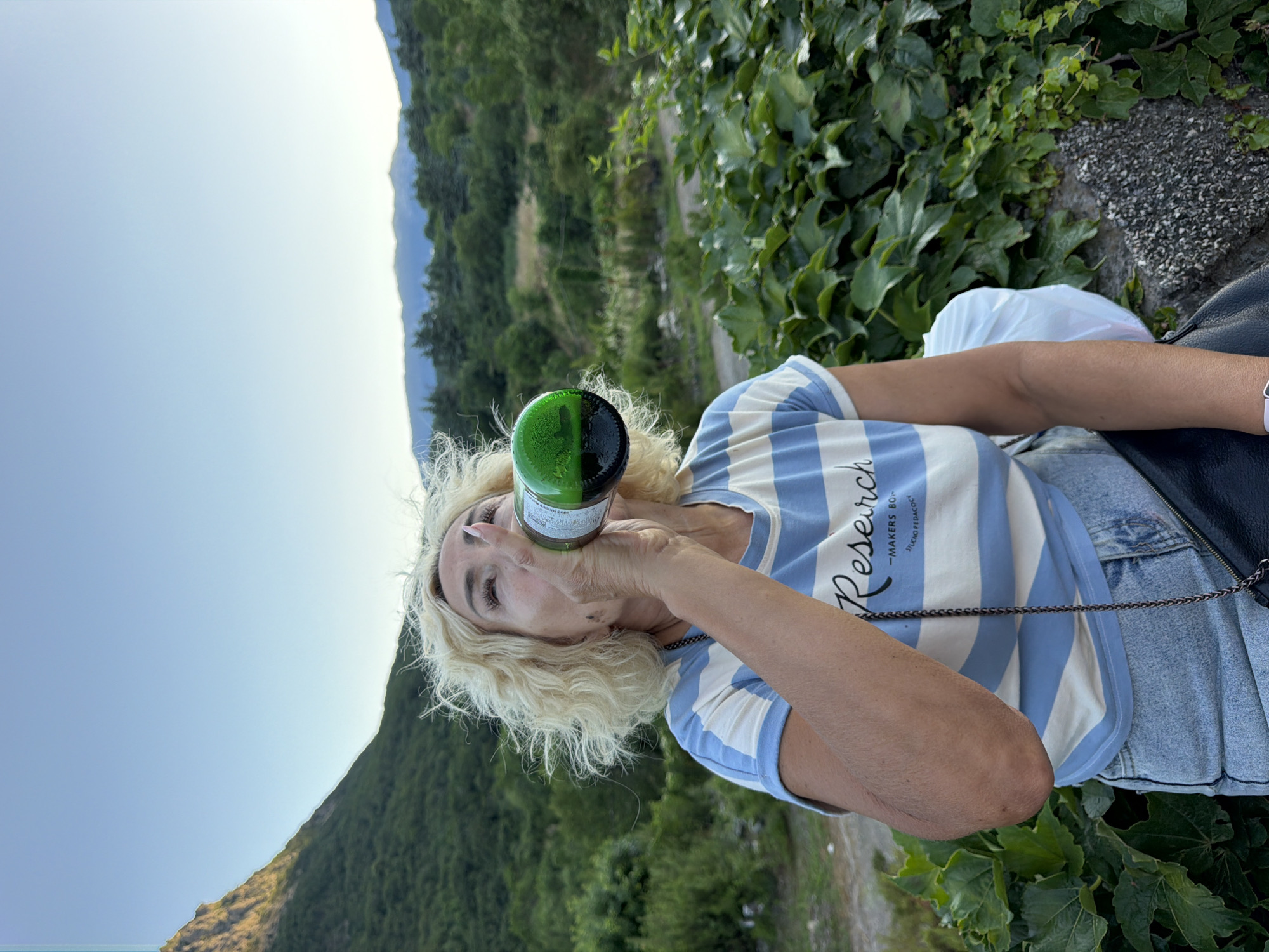 This is a vertical, medium shot of a woman with blonde, curly hair drinking from a green glass bottle outdoors. The woman is wearing a blue and white striped t-shirt and denim shorts. She is holding the bottle to her lips and appears to be drinking. Her eyes are closed and her head is tilted back slightly.
The background consists of lush green foliage and a distant mountain range under a clear, bright sky. The vegetation appears to be on a hillside, suggesting a rural or natural setting. The lighting indicates it is daytime, likely sunny.
The t-shirt has text on it that appears to read "Research -MAKERS BOD-". There is also a black bag with a chain strap visible on the woman's right side. The overall mood appears relaxed and informal. The presence of dense green growth and mountainous terrain hints at a location like the one described.