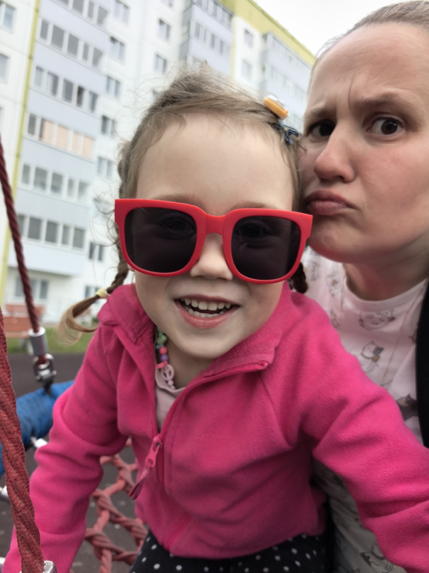A close-up, selfie-style photograph captures two females, likely mother and child, on a playground. The child, in the foreground, wears oversized red sunglasses and a bright pink hooded jacket over a light-colored shirt with a peace sign pendant. Her hair is in pigtails, one with a yellow tie and a small orange clip. The woman in the background, her face contorted in a playful pout, wears a white t-shirt with a repeated cartoon cat pattern and a black jacket or strap. They are positioned in front of a light-colored, multi-story residential building with numerous windows, characteristic of urban housing in cities like Froly, Russia. The setting appears to be an outdoor playground, with a red rope element of a swing or climbing structure visible on the left and a net structure behind the child. The lighting suggests an overcast day.