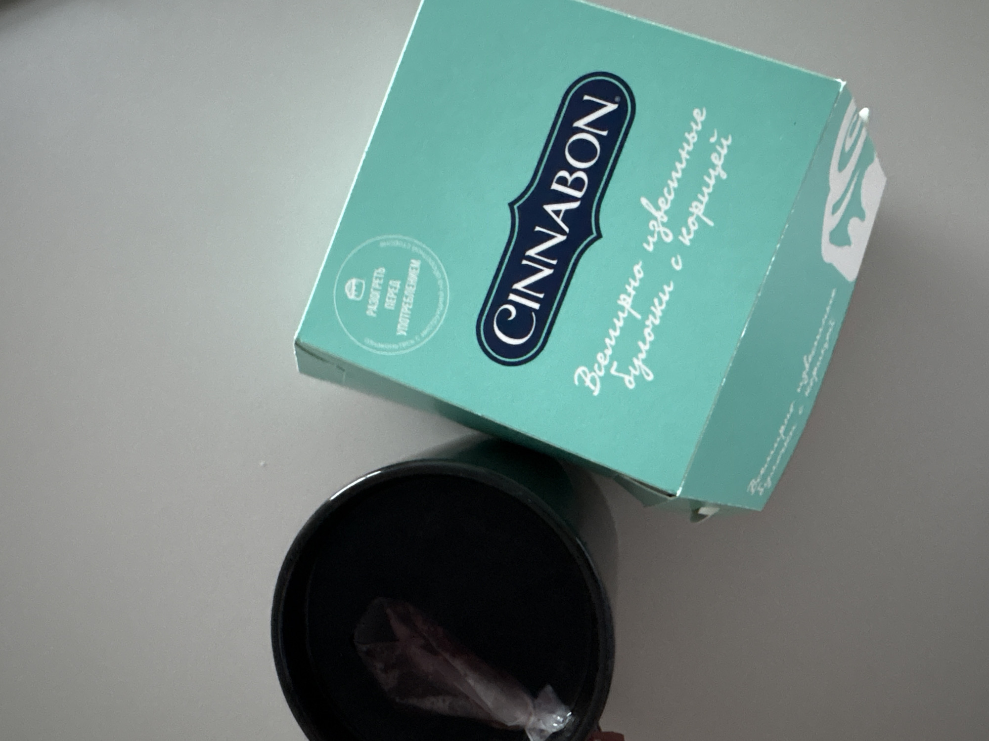 The image shows a box of Cinnabon cinnamon rolls and a black container. The box is light blue and has the Cinnabon logo and some Russian text that translates to "The world's most famous cinnamon rolls". The black container appears to contain an object wrapped in plastic. The items are situated on a gray surface.