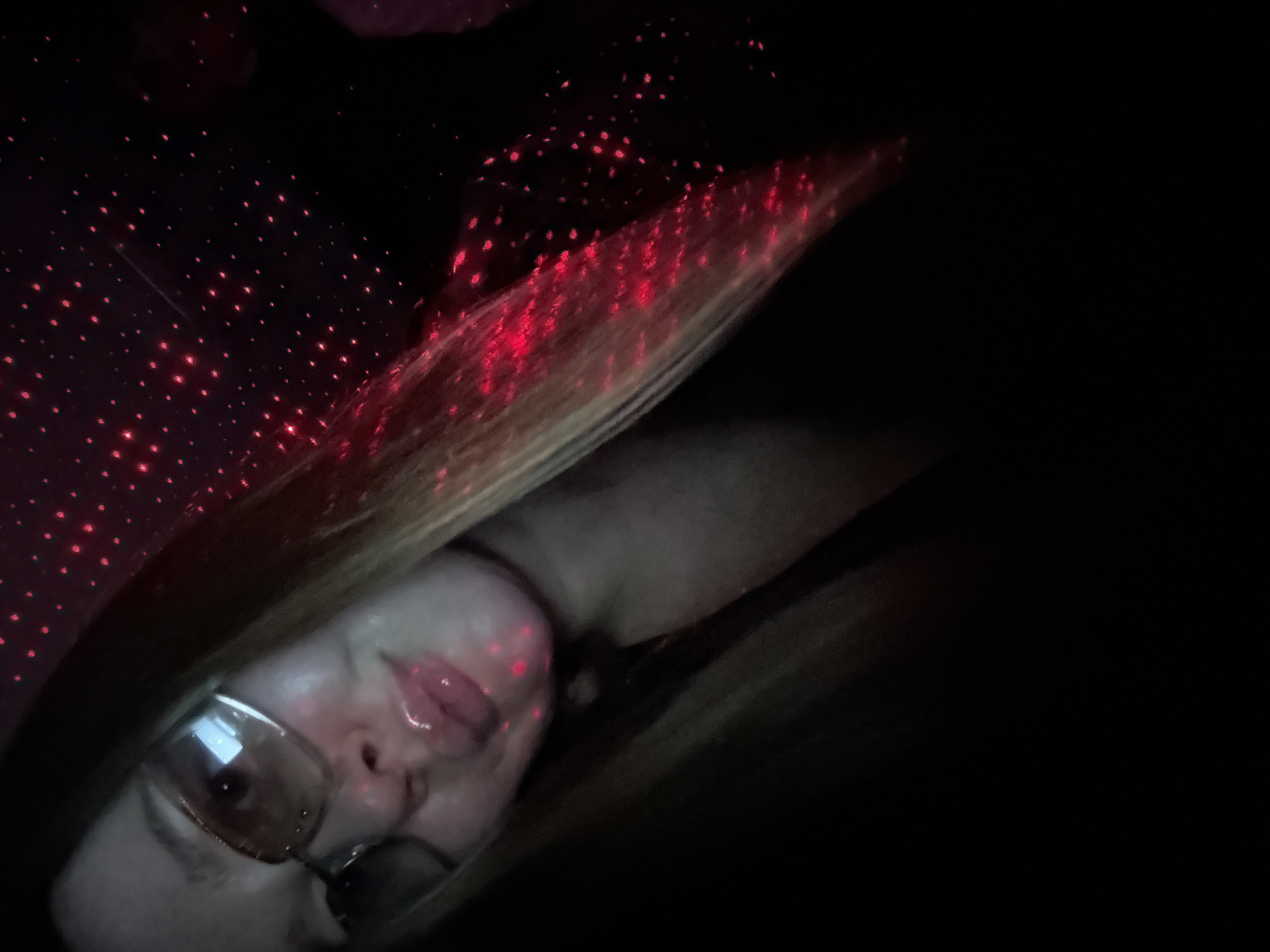 A selfie is taken by a young person wearing sunglasses, looking down at the camera. The person's lips are shiny and appear to be glossed. Red laser dots are projected onto the person's face, hair, and clothing, creating a speckled effect. The background is dark with more red laser dots scattered across it, suggesting the photo was taken in a dimly lit environment, possibly with a laser projector. The location is identified as Noviy, Russia.