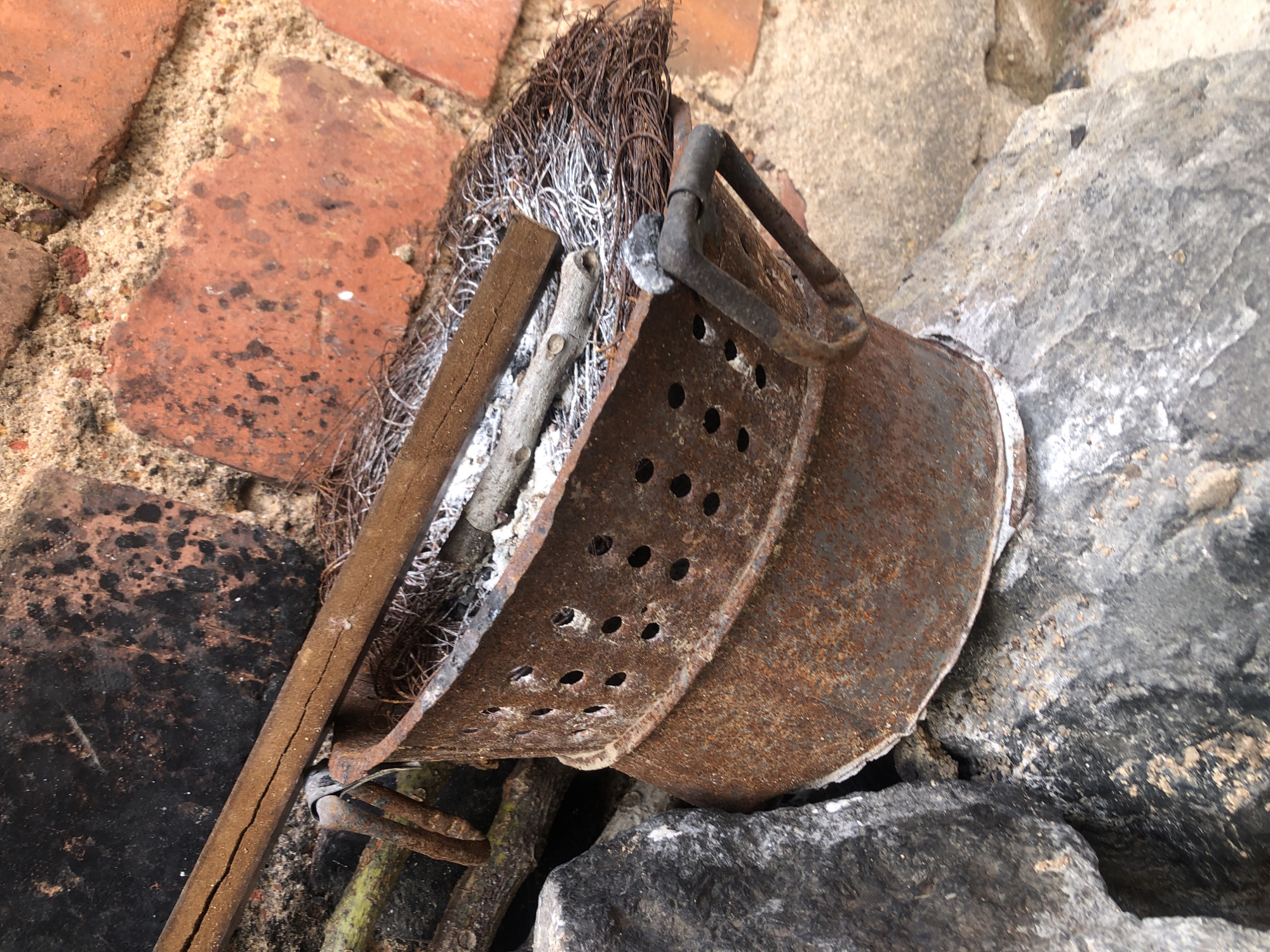 The image displays a weathered, rusty, perforated metal container, resembling a bucket or sieve, positioned on irregular gray and black stones. The container is heavily corroded, showing a dominant reddish-brown rust texture, and features multiple circular holes punched into its side. A dark, possibly metal, handle is visible on the right side. Inside the container are remnants of burnt material, including white ash, a short, light-gray wooden stick with small branches removed, and a darker, rectangular piece of wood exhibiting a central crack. A mass of thin, dark, wiry material, possibly burnt metal fibers or similar residue, is also present within and spilling slightly from the container.
Behind the container, on the upper right, is a wall constructed from reddish-brown bricks set with sandy mortar. Some bricks show dark discoloration or charring. To the upper left, a dark, textured surface, possibly a charred wall section or another background element, is visible. The entire scene is illuminated by natural light, highlighting the textures of the rusted metal, stones, and brickwork. No people, specific actions, or direct interactions are visible. The location is in the city of Unknown, Unknown.