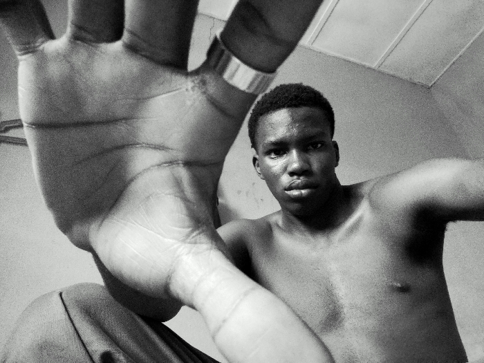 A monochrome, low-angle photograph depicts a shirtless adult male, likely of African descent, situated in an indoor location in Keffi, Nigeria. His left hand is extended towards the camera in the immediate foreground, palm open and fingers splayed, occupying the left side of the frame. A metallic ring is visible on the ring finger of this extended hand. The individual gazes directly into the camera with a neutral expression; his face and upper torso exhibit a discernible sheen. A portion of his lower body, covered by a draped fabric, is visible in the lower left corner. The background consists of a plain, light-toned wall and a ceiling featuring a rectangular panel or fixture in the upper right.