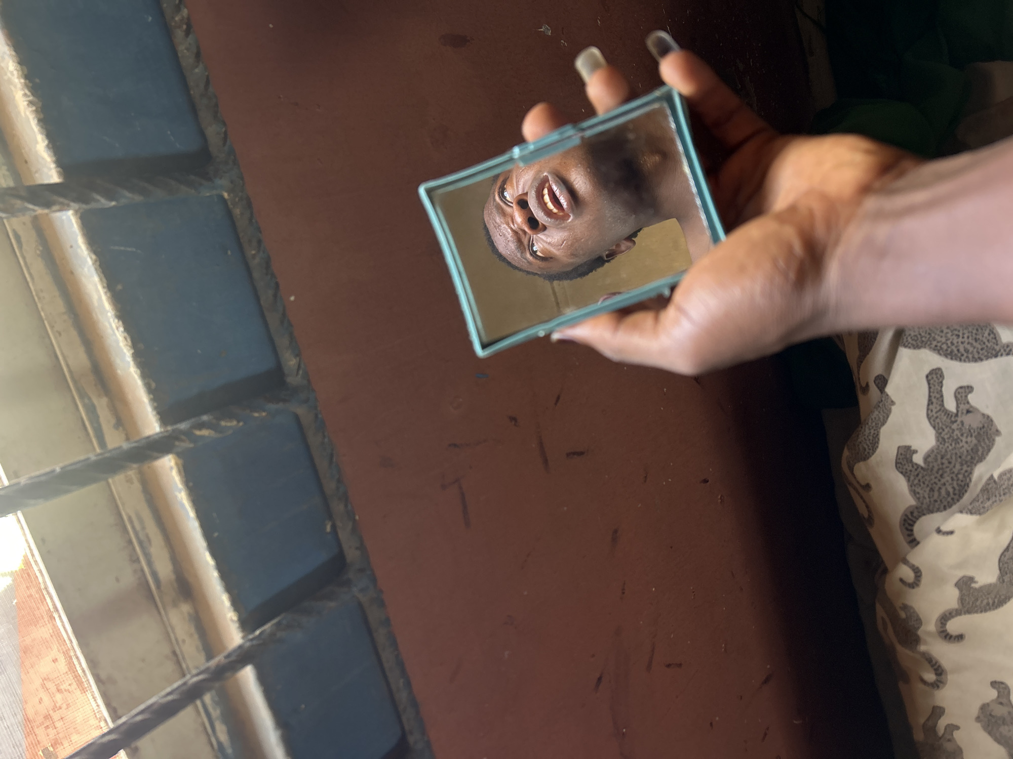 The image captures a close-up view of a person's hand holding a small, rectangular mirror with a light blue frame. The mirror reflects the expressive face of a person with dark skin, seen at an angle. The reflected face has an open mouth, revealing teeth, and eyes that appear to be looking upwards or rolled back, conveying an emotion that could be surprise, laughter, or playful joy. The hand holding the mirror has long fingernails, some of which are painted a dark color.
In the background, to the left, there is a section of what appears to be a window or a vent, characterized by horizontal metal bars. Some of these bars are painted a muted blue or grey, and light passes through them, suggesting it is daytime. The prominent surface behind the hand and mirror is a reddish-brown or maroon wall or floor, showing some texture and minor marks. To the right, a patterned fabric is visible, possibly part of clothing or bedding, featuring grey, stylized animal figures resembling leopards or jaguars on a light cream background. There is no visible text in the image. The setting appears to be indoors, possibly a room with natural light.