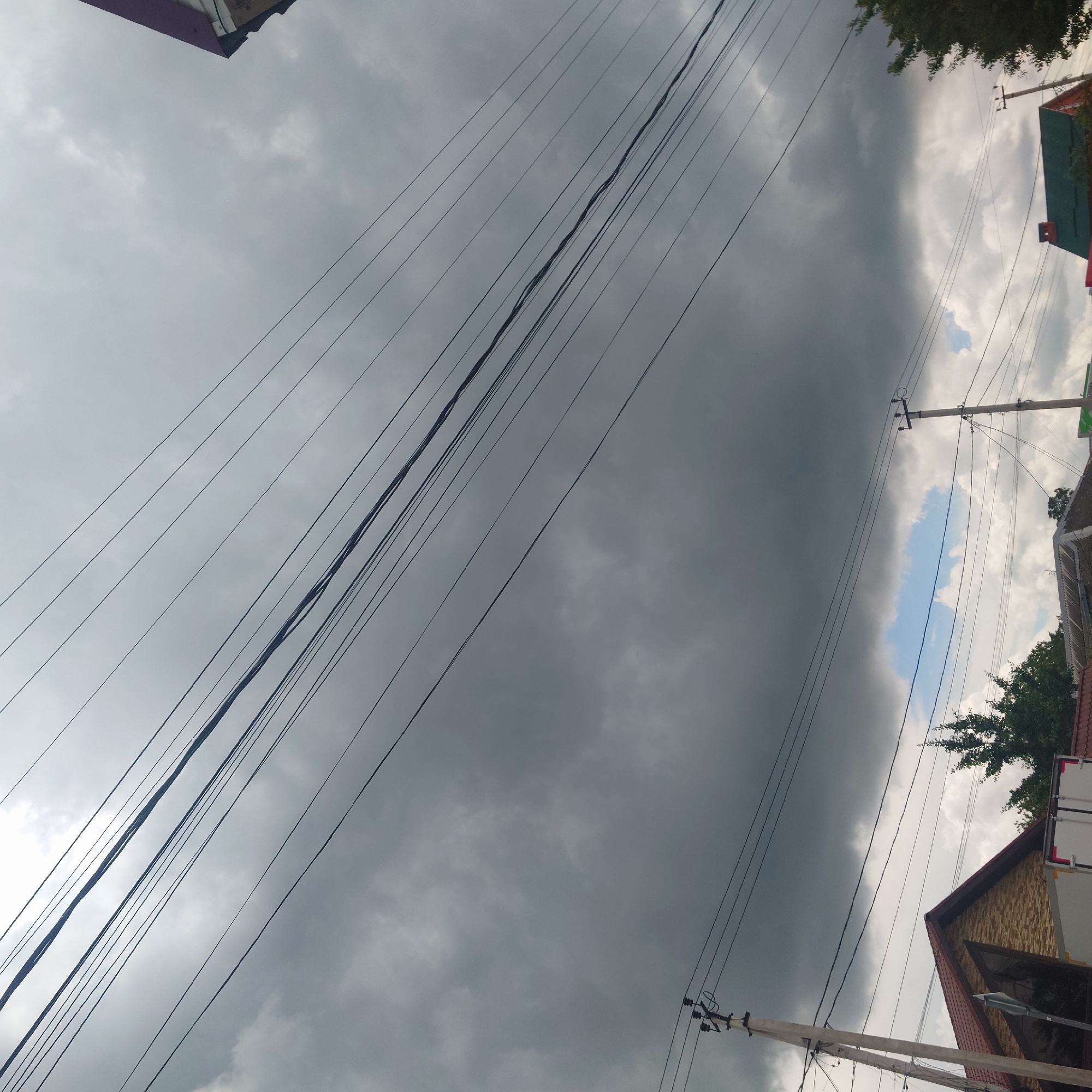 The image depicts an outdoor scene, likely in a residential area, with a focus on utility lines and overcast skies. The sky is dominated by dark, heavy clouds, suggesting impending rain or a storm. Several parallel utility wires crisscross the frame, running diagonally from the top left towards the bottom right.
At the bottom of the image, parts of houses and buildings are visible. On the left, a portion of a house with a brown, textured facade and a dark-pitched roof is partially in frame. A light-colored utility pole stands to its left, supporting some of the overhead wires. Further to the right, another utility pole is visible, as is a section of a building with a green roof and a red brick chimney. Trees with dense green foliage are scattered throughout the lower portion of the image, some appearing mature and others younger. A sliver of bright blue sky with some white clouds is visible between the darker cloud formations.
The scene is devoid of any visible people or active subjects. The overall impression is one of an ordinary urban or suburban environment under a turbulent sky, with the infrastructure for power and communication being a prominent feature. The location is identified as Belorechensk, Russia.