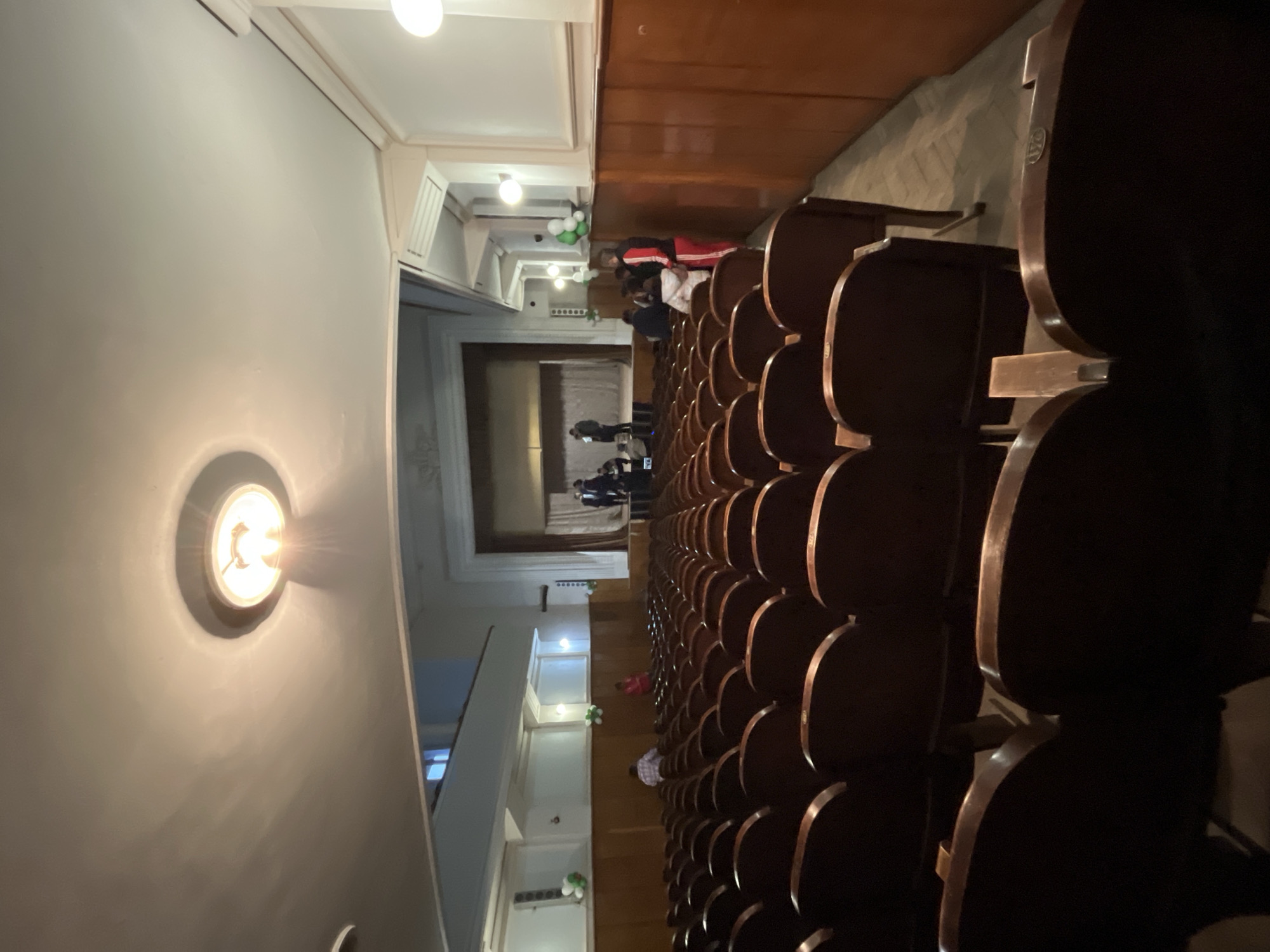 The image shows the interior of a theater or auditorium, oriented vertically. Rows of empty dark brown or black seats fill the right side of the frame, suggesting a large seating capacity. People are visible in the background toward the stage area, some standing and some sitting. The stage itself appears to be covered by a curtain. The left side of the image shows a wall with a circular light fixture illuminated, emitting a warm glow. The environment appears to be somewhat old-fashioned, with classic architectural details. It is not possible to determine the location within Voiniagovo, Bulgaria from this image.