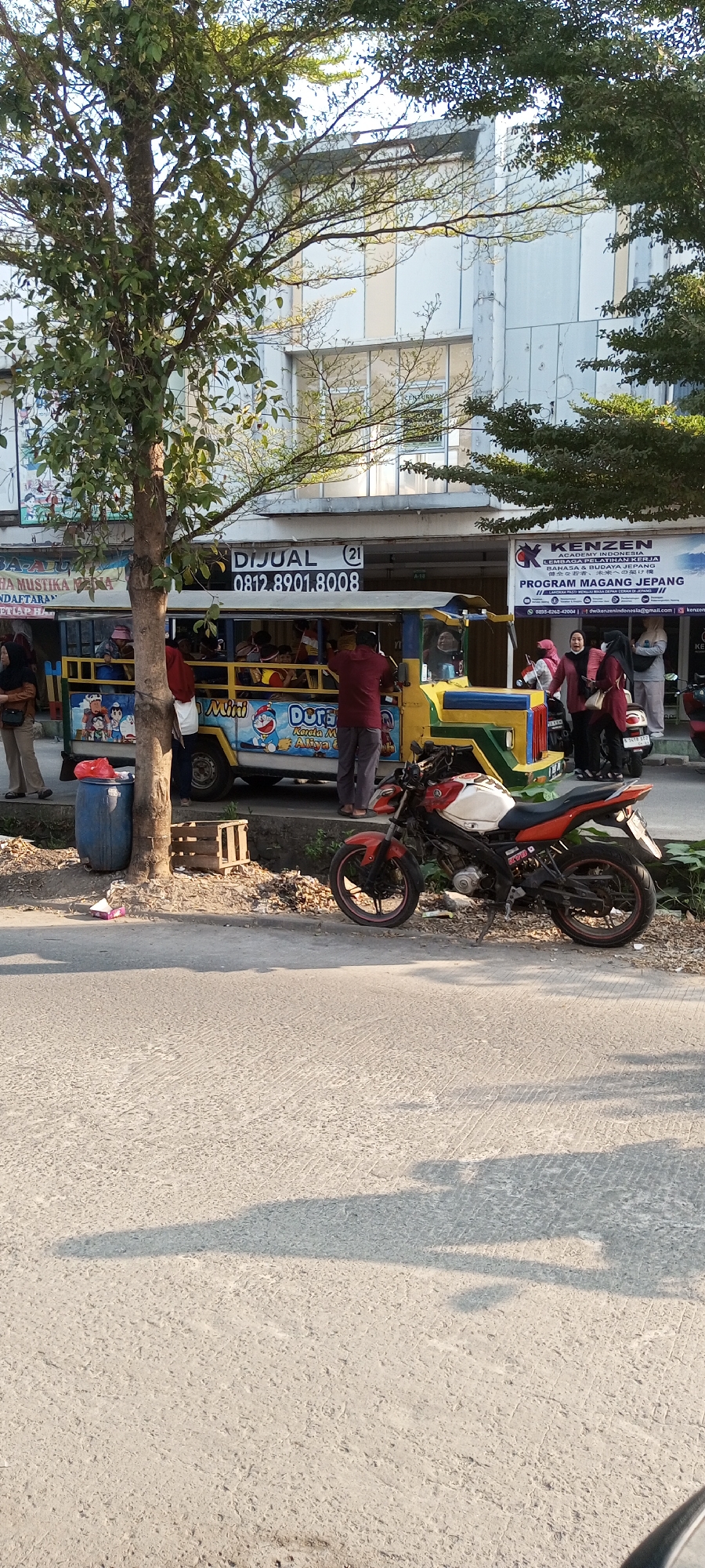 In Setu, Indonesia, on a sunny day, a red and white motorcycle is parked on the side of a road. Behind it, a yellow and blue open-air passenger vehicle, decorated with cartoon characters like Doraemon, is stopped. Several people are visible in and around the vehicle, some standing, others seated. A blue barrel and a wooden crate are near the vehicle. In the background, a modern building with large windows stands, with trees providing shade in the foreground. Signs are visible on the building and the passenger vehicle, including one that says "DIJUAL" (For Sale) with a phone number.