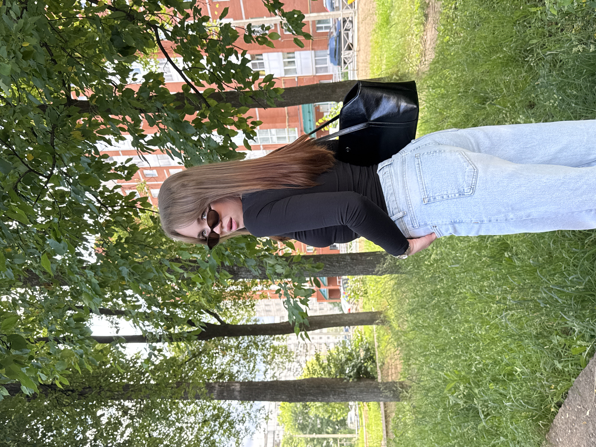 In Chaikovsky, Russia, a young woman with blonde hair is captured from the waist up, turned away from the camera and looking over her left shoulder. She wears a black long-sleeved shirt, light blue jeans, and dark-rimmed sunglasses. A black purse hangs from her right shoulder. She is standing in an outdoor setting with a patch of grass and trees in the foreground and a brick building in the background. The lighting suggests it is daytime.