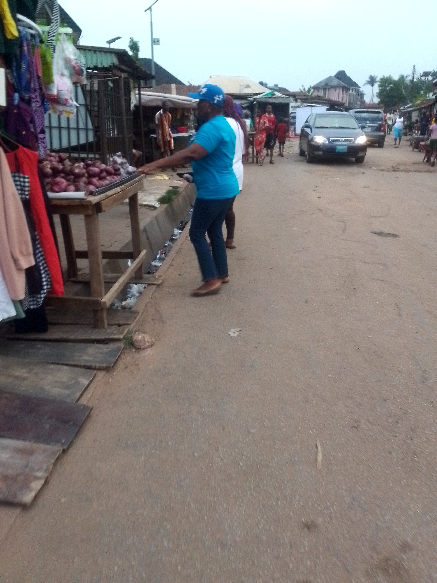 The image shows a street scene in Onitsha, Nigeria. In the foreground, a man wearing a blue shirt and hat is standing next to a wooden table displaying piles of red onions. A rack of clothes is hanging to the left of the table. A second person is partially visible behind the man. In the background, there are several people walking along the street and two cars are visible on the right-hand side. The street is unpaved and buildings line either side.