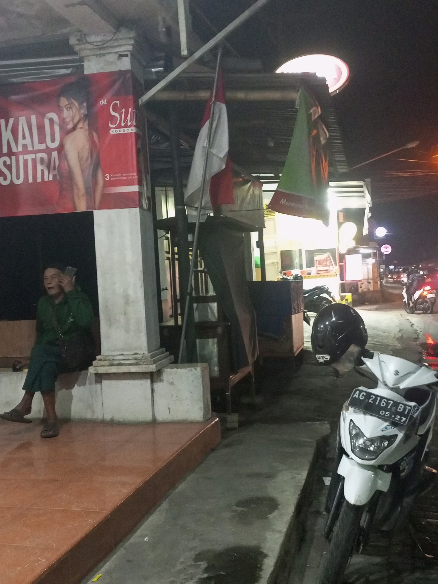 A medium shot captures a scene at night in front of a building, possibly a shop or business, in the city of Ngasem, Indonesia. A banner with the text "KALO SUTRA" and an image of a woman is prominently displayed on the left. To the right of the banner, an Indonesian flag is partially visible.
A person, appearing to be an adult, is seated on a low wall on the left side of the frame, wearing a green jacket and pants, and is holding a mobile phone to their ear, seemingly engaged in a conversation. They are wearing sandals. In the foreground, a white scooter is parked, facing forward, with a license plate that reads "AC 2167 BT 05-27". A black helmet is placed on the ground near the scooter. The ground appears wet in places, suggesting recent rain or dampness. The background shows illuminated signs and structures typical of a commercial area at night. The overall lighting is dim, with artificial lights providing illumination.
