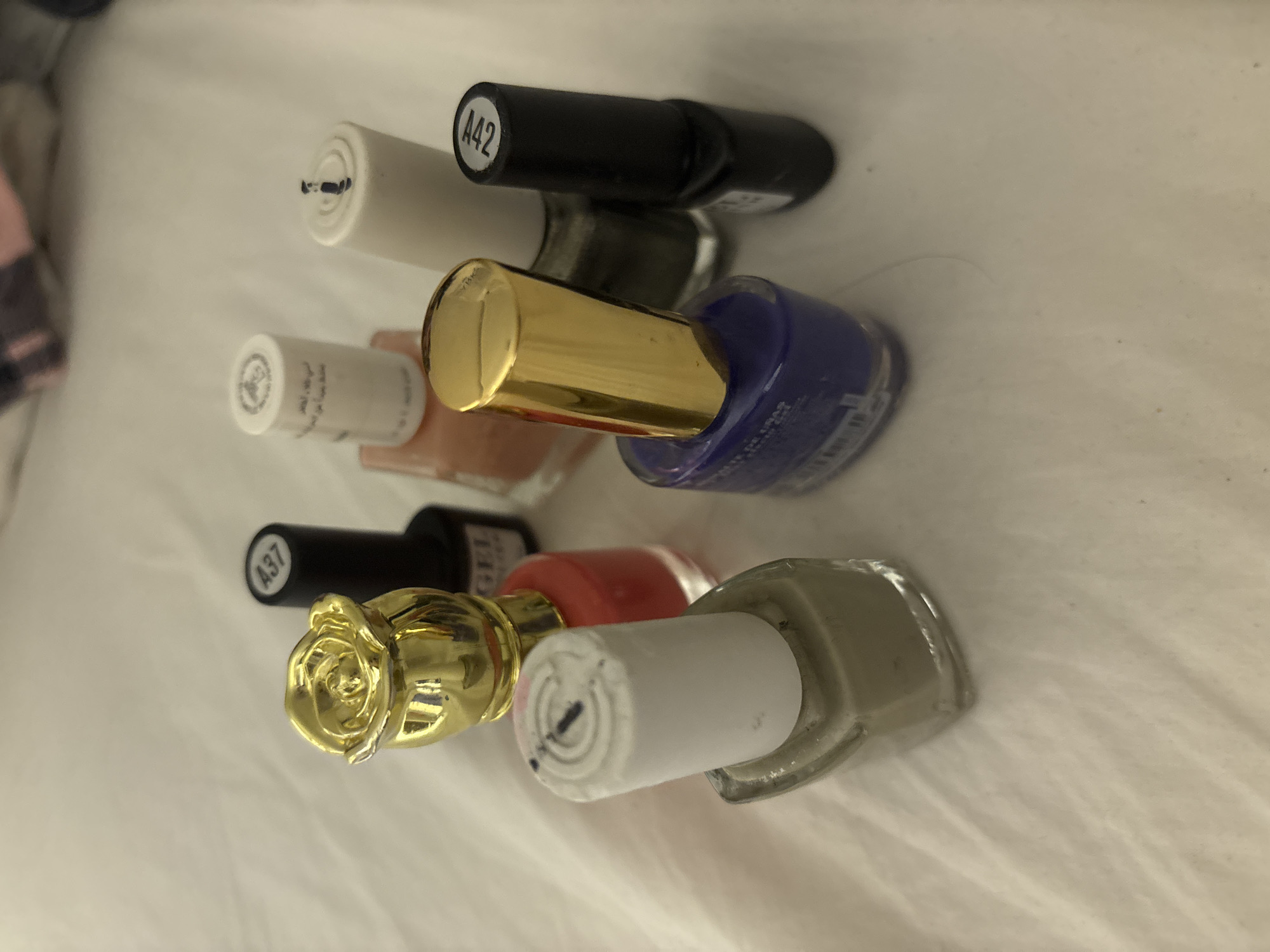 A collection of nail polish bottles are arranged on a white, textured surface, possibly a bedsheet. The bottles vary in color, shape, and design. One bottle has a gold rose-shaped lid, another a gold cylindrical lid, and others have plain white or black caps. Some caps have labels with alphanumeric codes such as "A37" and "A42". The nail polish colors visible include red, light green, purple, and a peachy-pink. The lighting is soft and diffused, suggesting an indoor setting. There are no people or indications of a specific location within Dubai, United Arab Emirates.