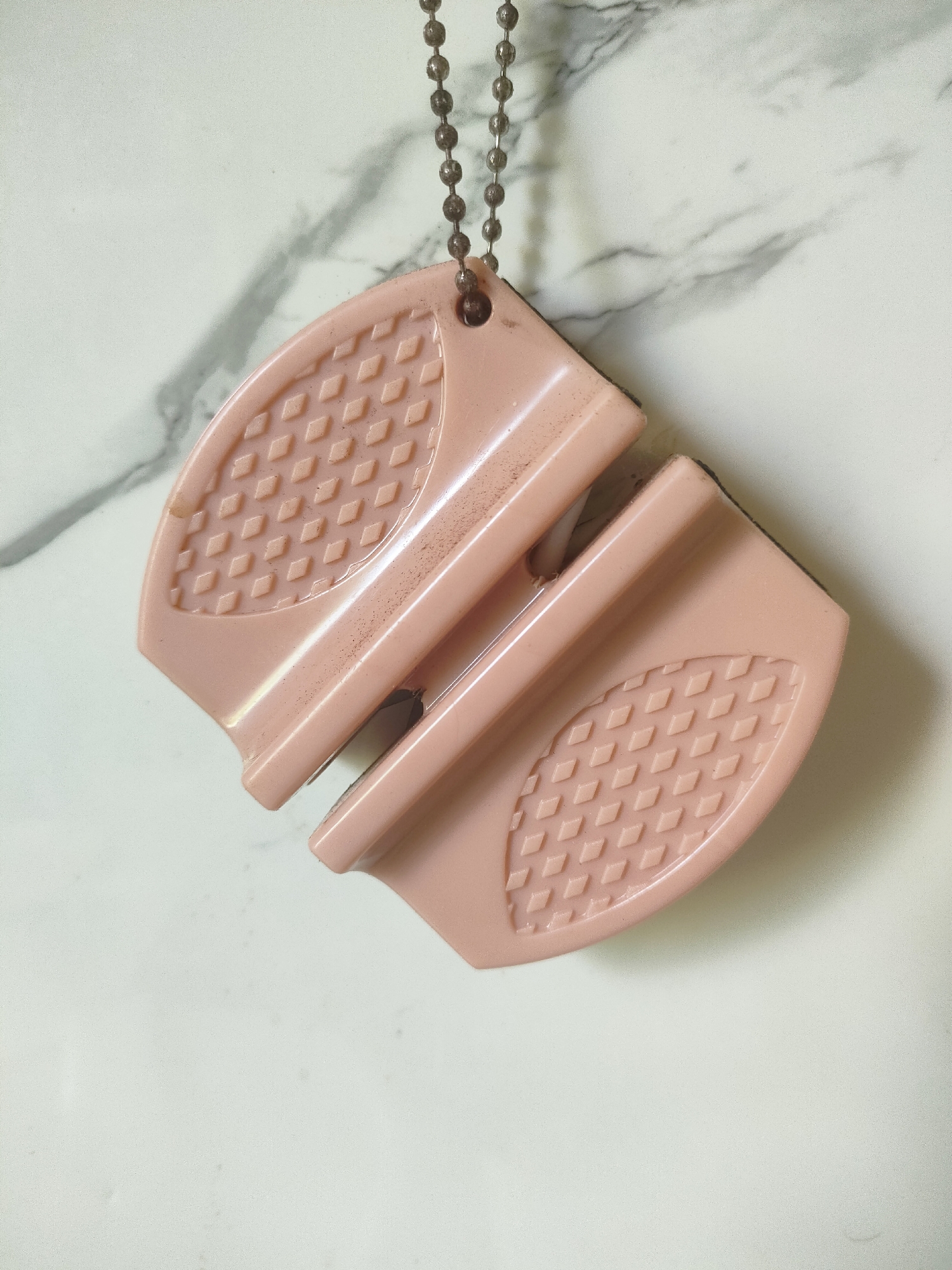 This media file features a close-up, overhead shot of a light pink, plastic object with a beaded chain attached, resting on a marble-patterned surface. The object appears to be a tool, possibly for cleaning or grooming, with textured diamond patterns on its curved ends and a slotted central section. The surface beneath is a light gray and white marble print. There are no people visible in the image, nor any discernible activity taking place. The lighting suggests an indoor setting, possibly a countertop or table, with no indication of the time of day or weather. The overall impression is of a simple, everyday item photographed against a neutral background. The object shows some minor dust or smudges, suggesting it may have been used. No text is visible in the image.