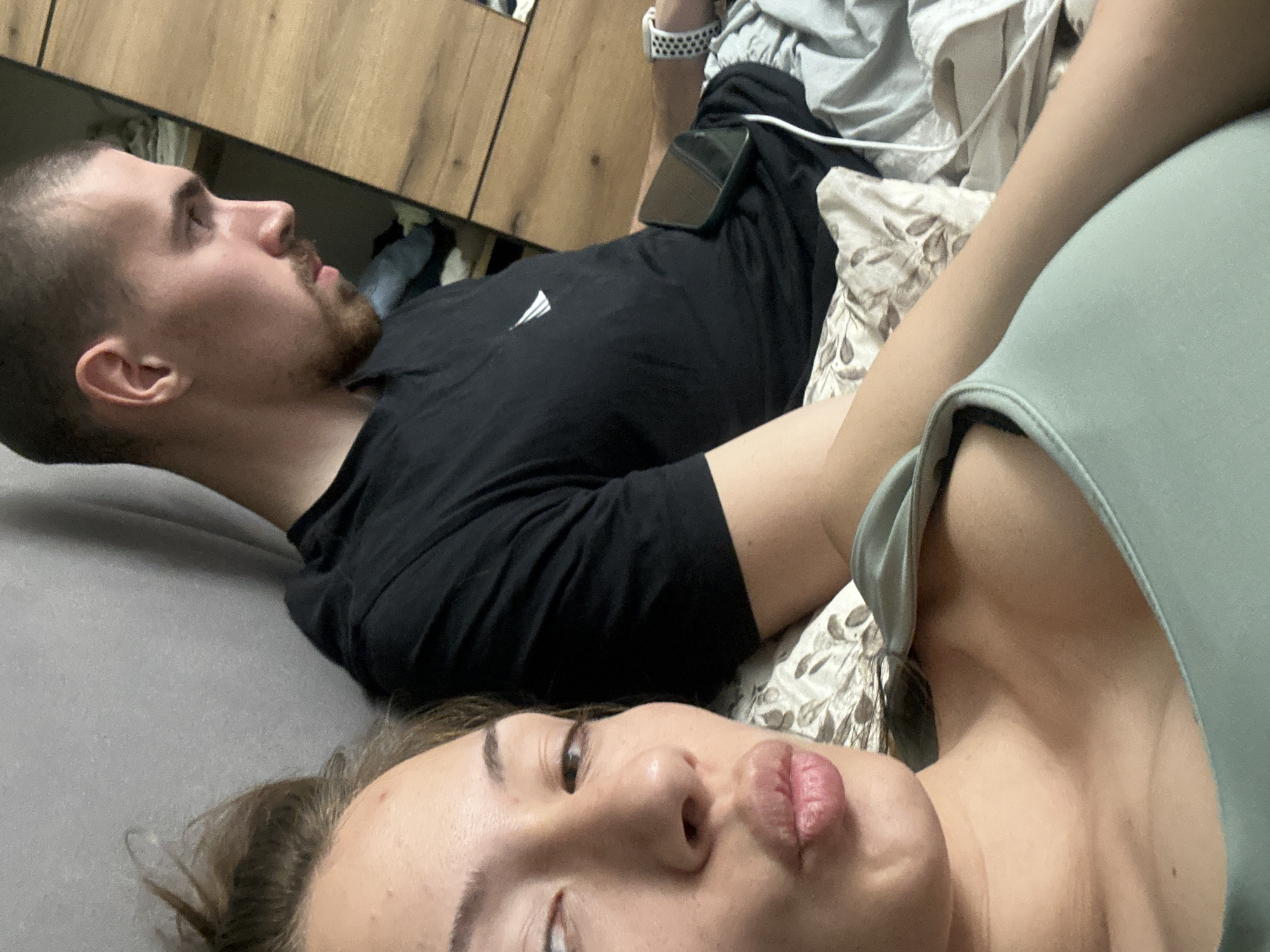 A couple is lying on a bed. The man is facing left with a beard and short hair, wearing a black shirt. A black smartphone rests on his thigh. The woman is closer to the camera, facing upwards, with her lips pursed. She wears a light green top. The bed is covered in grey sheets and a patterned blanket. The background shows a wooden cabinet. The location is unknown, not necessarily Burgas, Bulgaria.