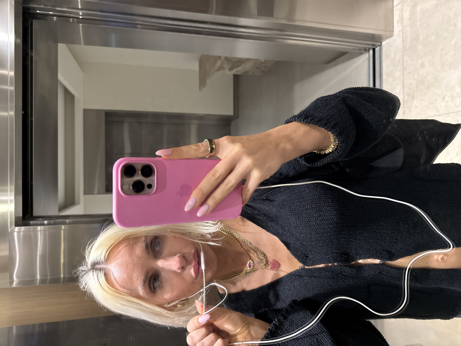 A blonde woman in a black knit sweater takes a selfie in front of a stainless steel surface, likely an appliance. She holds a pink iPhone case. She wears layered gold necklaces and a ring on her right index finger and a tennis bracelet on her right wrist. White earphones are in her ears. Reflected in the surface are white walls and stainless steel appliances. The floor appears to be beige tile.