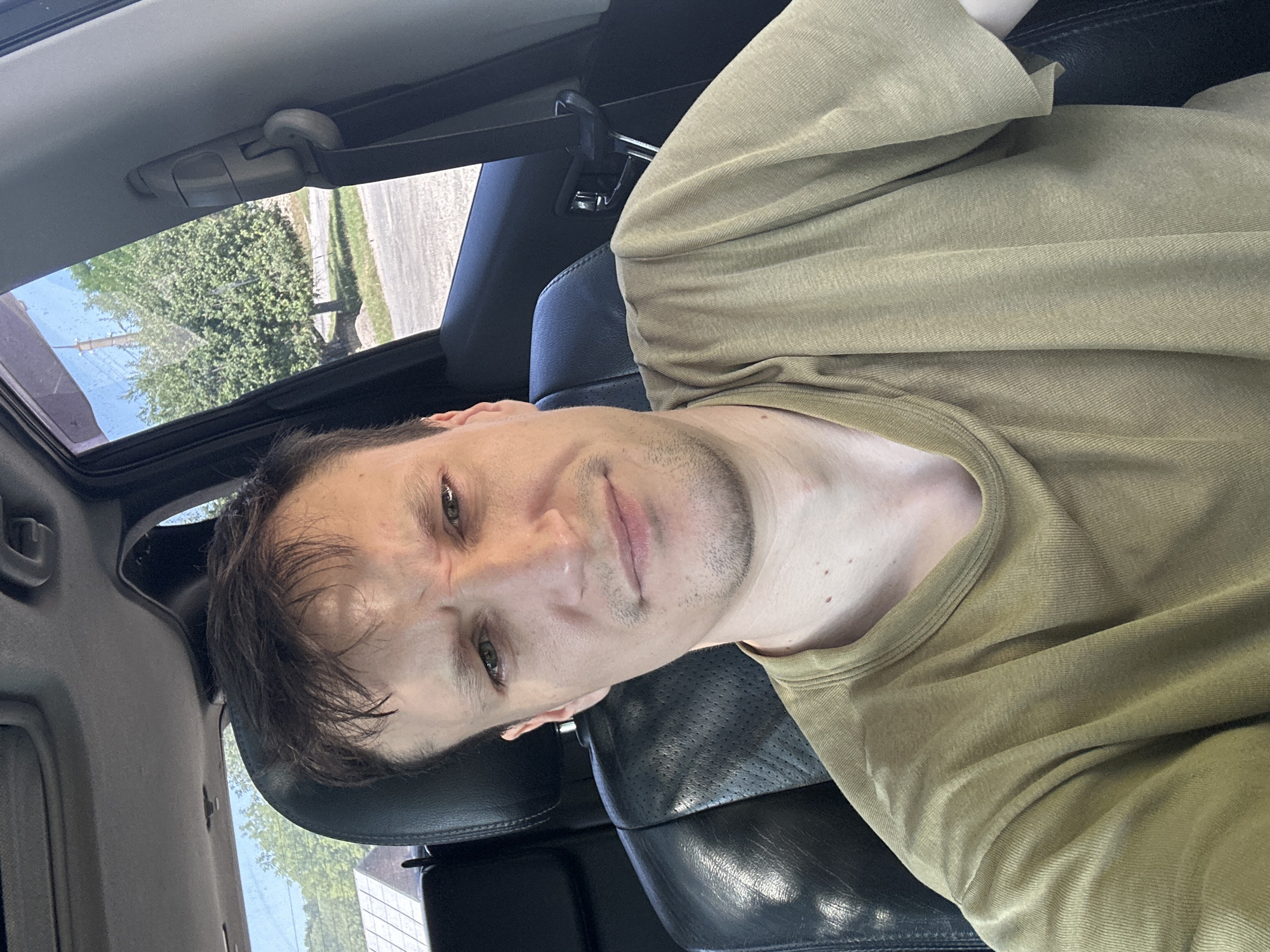 The image shows a man sitting in the driver's seat of a car. He is wearing a light brown t-shirt. The car's interior is visible, including the black leather seats and part of the dashboard. Through the car window, trees and a building are visible, suggesting an outdoor location. The man is looking at the camera with a neutral expression. The image appears to be a selfie taken from the perspective of the driver. There is no specific information about the location in the city of Makiv, Ukraine within the image itself.