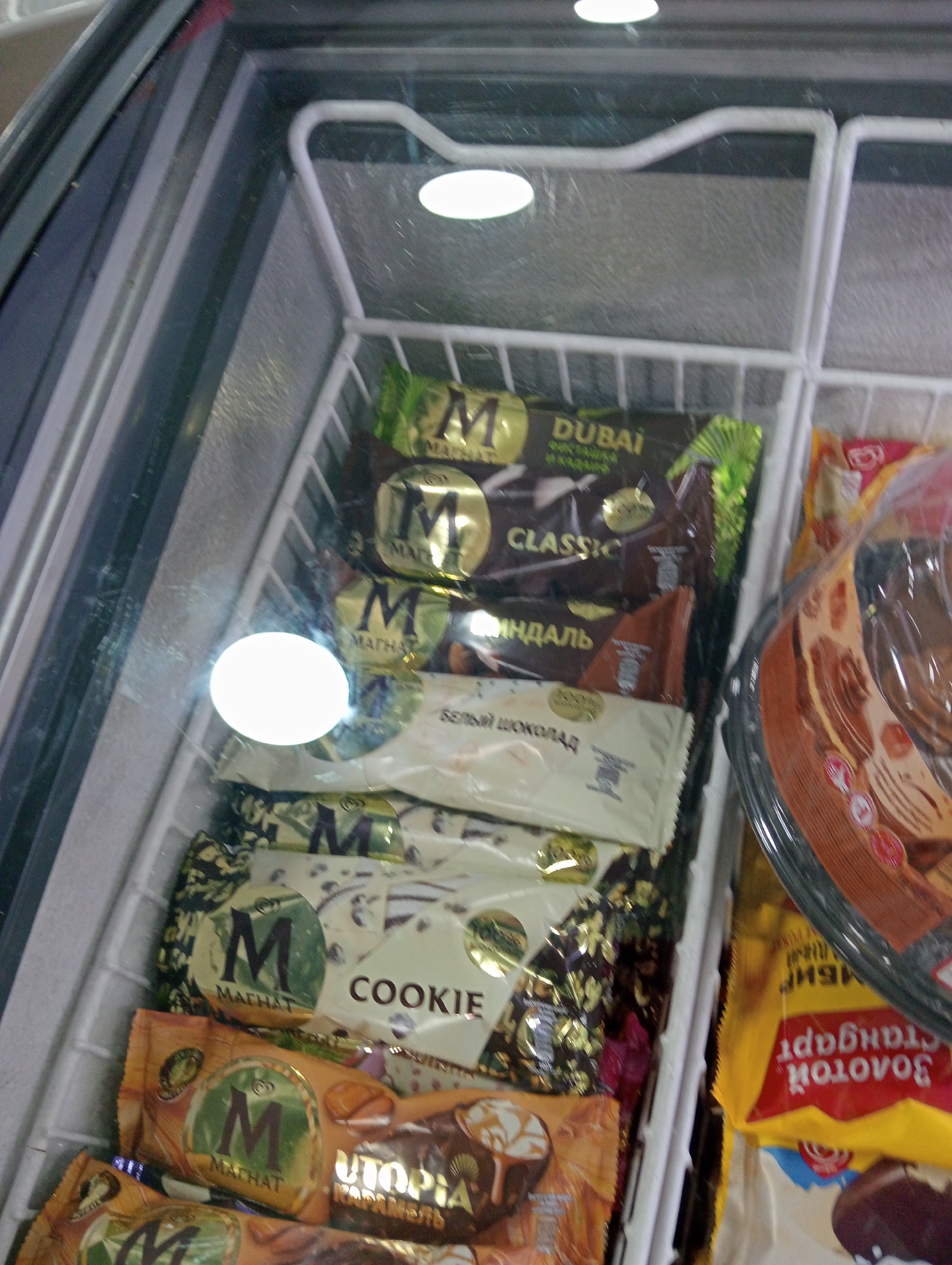 The image shows an interior view of a freezer chest with multiple rows of individually wrapped ice cream bars. The ice cream bars are predominantly packaged in foil wrappers with various colors and branding. Some wrappers feature the "M" logo and text such as "DUBAI," "CLASSIC," "COOKIE," and "UTOPIA." There are also some ice cream products in transparent plastic packaging that appear to be tubs or cakes. The freezer interior is white, and a white wire rack system is visible holding the products. Overhead lighting casts reflections on the freezer door. The overall environment suggests a retail setting, such as a convenience store or supermarket.