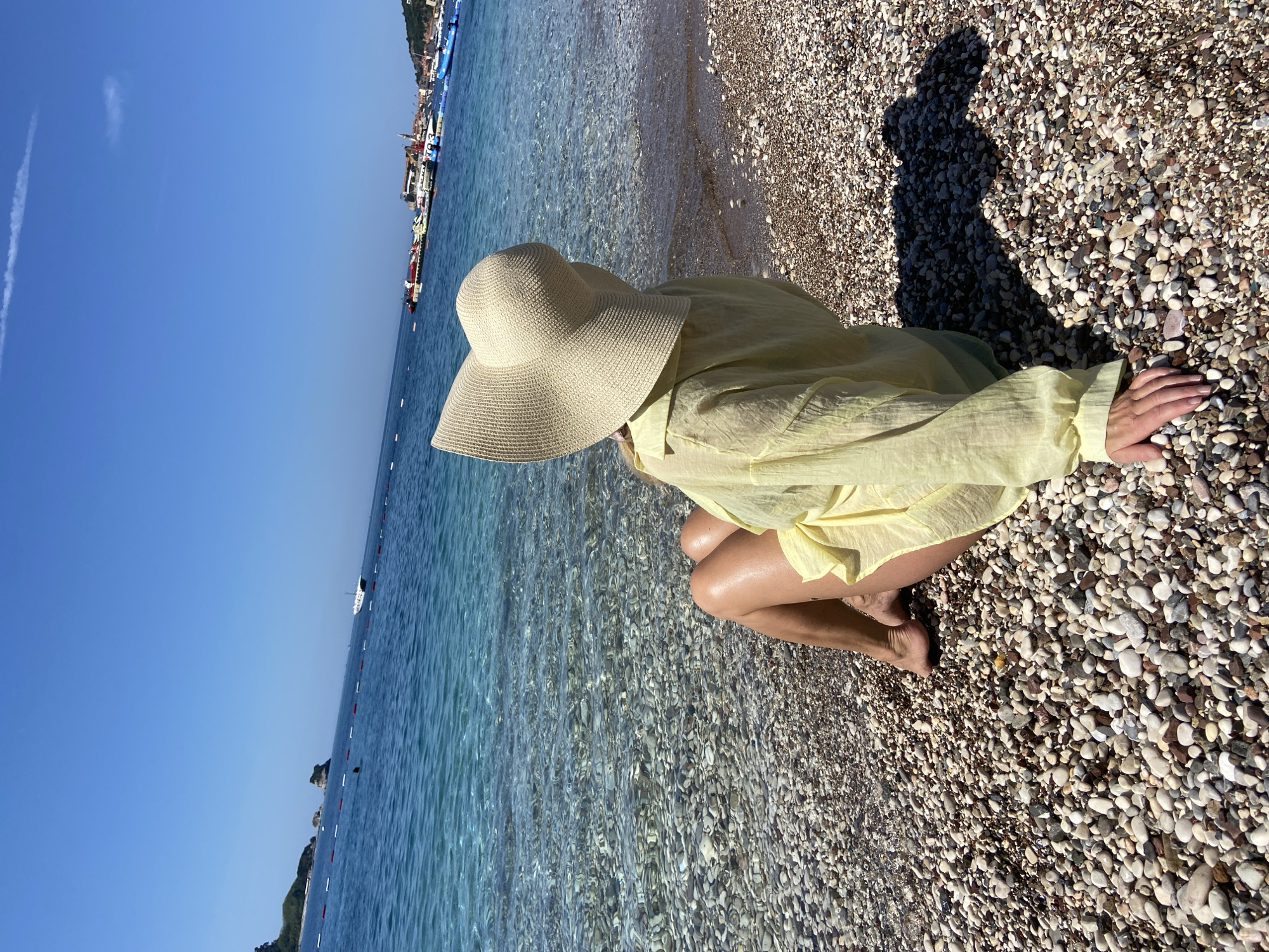 The image captures a person sitting on a pebbly beach under a clear, bright blue sky, gazing out at the Adriatic Sea in Budva, Montenegro. The individual, viewed from behind, wears a light yellow long-sleeved cover-up and a wide-brimmed, light-colored straw hat. Their left hand rests on the multi-colored pebbles of the beach, which transition into exceptionally clear, shallow turquoise water, revealing the stones beneath. Further out, the sea deepens to a rich blue, with a visible line of red and white buoys marking a boundary.
In the left background, rocky, vegetated coastline and two distinct rocky islets emerge from the sea. A white boat is visible navigating beyond the buoy line. The right background features the historic Budva Old Town, characterized by its cluster of buildings with distinctive red-tiled roofs and white facades, nestled against a hillside. A busy marina extends from the town, filled with various boats, pontoons, and water activity equipment. The entire scene is bathed in bright sunlight, casting distinct shadows on the pebbles. A faint contrail streaks across the otherwise cloudless sky.