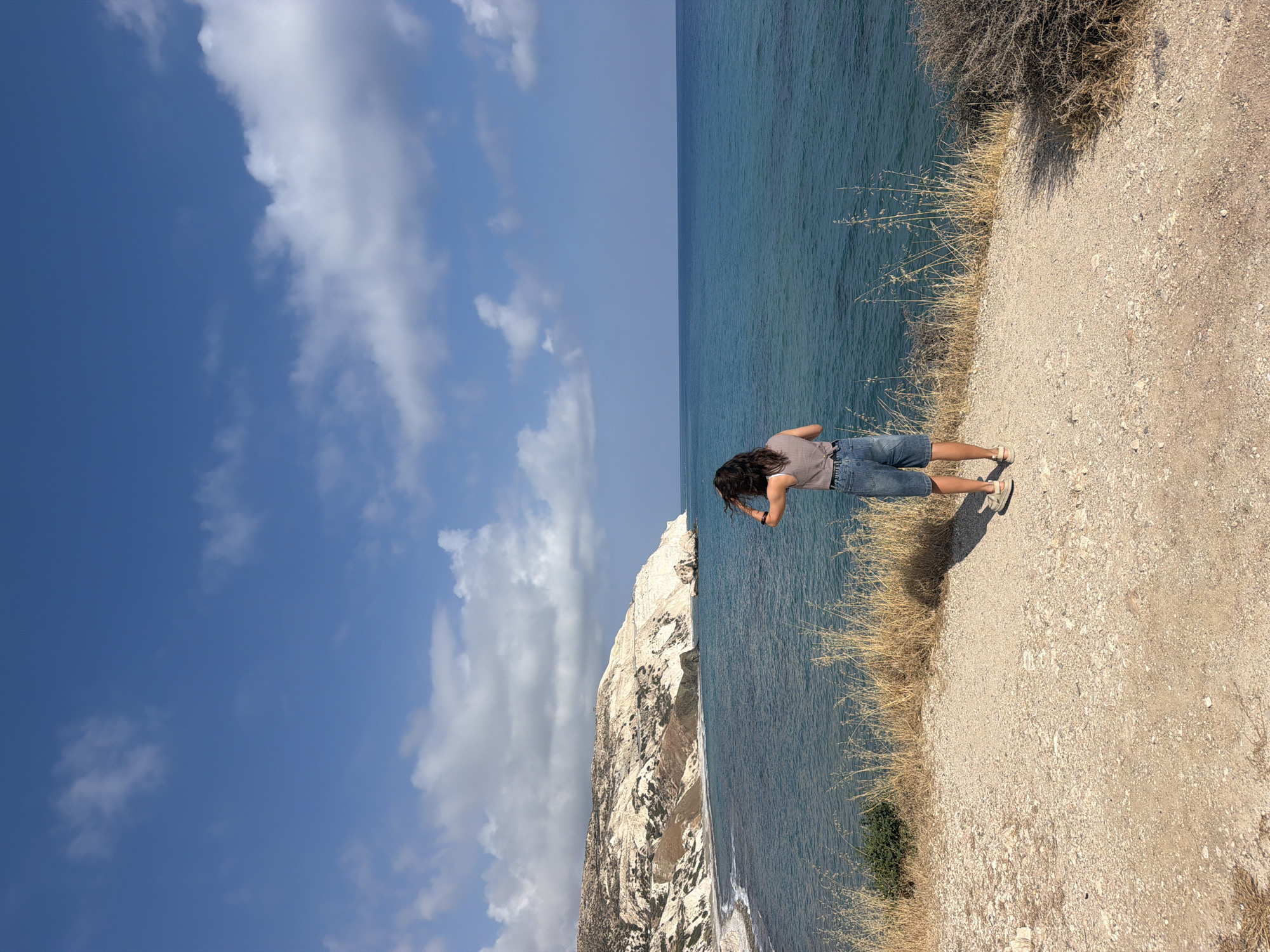 A person stands on a dirt path overlooking a turquoise sea. The person, viewed from behind, has dark, curly hair and wears a grey t-shirt and denim shorts. They are touching their hair with their right hand. The path is covered in small stones and dry grass grows along its edge. In the distance, a white, rocky cliff face juts into the sea. The sky above is a bright blue with scattered white clouds.
