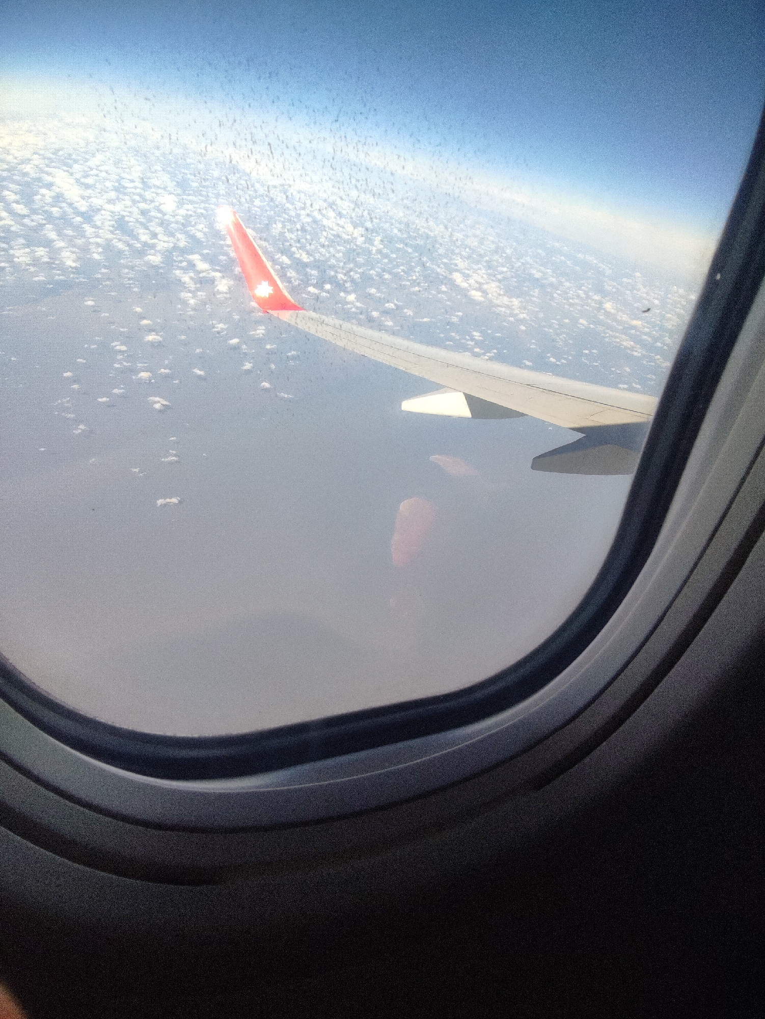 The image shows an airplane wing and clouds seen through an airplane window. The wing has a red tip. The environment appears to be high altitude. It is not possible to determine the location as Chaikovsky, Russia, from the image. No people are visible.