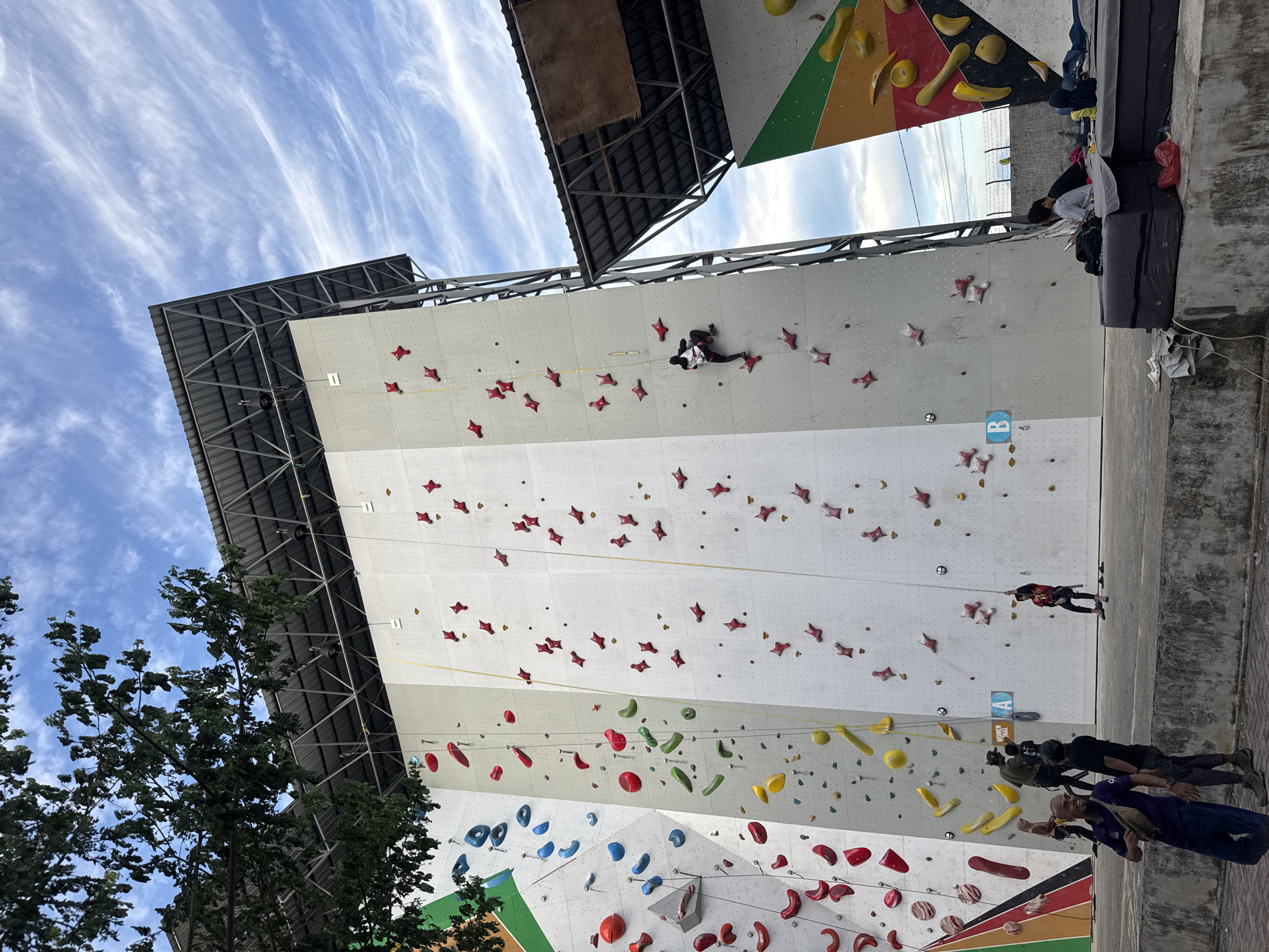 An outdoor climbing wall facility is depicted with multiple sections of varying difficulty and colors. The main wall is white with red and pink climbing holds arranged in a star pattern. To the left, another section of the wall features blue and red holds on a white background, with a multi-colored triangular section on the far left. A smaller, separate climbing wall structure is visible to the right, also with colorful climbing holds. The sky is partly cloudy with wispy white clouds against a blue background.
Several individuals are present. On the main white wall, one person is actively climbing. Near the base of this wall, a group of people are gathered, some appearing to be instructors or participants. Another individual is seen rappelling down the wall on the right side. At the bottom of the scene, a few people are sitting on crash pads in front of the climbing walls, and others are walking around. One person in the foreground is walking towards the camera, wearing a dark purple shirt and dark pants, with a light brown waist bag.
The facility appears to be in an urban or semi-urban environment, possibly a recreational area. Given the context of the user's request, the facility is identified as being in Banda Aceh, Indonesia.