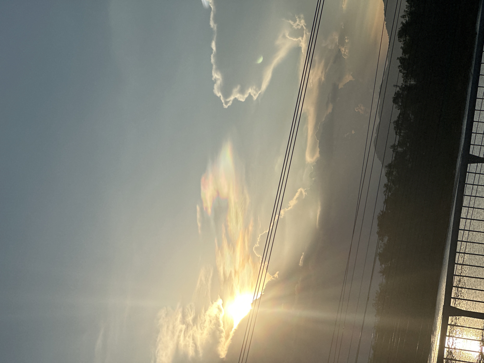 The image depicts a sunset in Nha Trang Ward, Vietnam. The sun is partially obscured by clouds, creating a burst of light rays. Power lines are visible against the sky. Silhouetted trees are on the right side of the frame, with a body of water reflecting the sunset. A portion of a bridge or railing is also visible on the right.