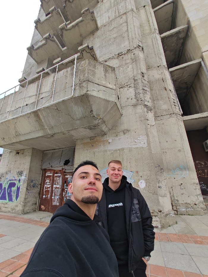 The image depicts two males posing for a selfie-style photograph in front of a large, multi-story concrete building located in Shumen, Bulgaria. The male in the foreground, positioned left, has dark hair, a short beard, and wears a black hoodie; he looks towards the camera with a neutral to slight smiling expression. The male in the background, positioned right, has short light hair, a smiling expression, and wears a black hoodie with a white graphic beneath a black jacket featuring subtle patterned sleeves.
The concrete building behind them is of brutalist design, characterized by prominent, irregular cantilevered balconies and platforms. The structure appears unfinished or abandoned, exhibiting significant disrepair including weathered and cracked concrete, spalling, and open apertures where windows would be. The ground floor features heavily rusted double doors, one visibly damaged, and sections of the lower concrete walls are covered with graffiti, including purple script. The immediate foreground shows a paved sidewalk composed of brown and lighter-colored tiles. The sky is overcast.