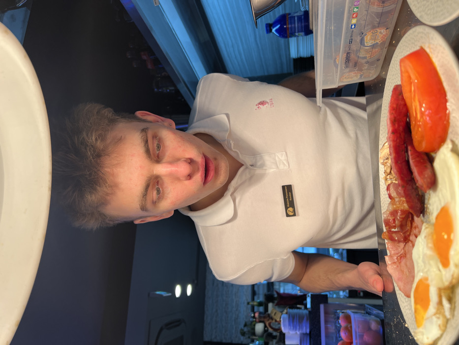 A young adult male, identified by a black name tag with a gold "Z" logo as Maksim Ponomarev, a waiter ("Konobar"), is captured leaning forward over a service counter. He wears a white U.S. Polo Assn. polo shirt. His eyes are directed downward, focused intently on a white plate situated on the counter. The plate holds a breakfast arrangement consisting of two fried eggs, strips of bacon, slices of ham, two reddish sausage links, and a single prominent red tomato slice. His mouth is slightly agape, indicating concentration or perhaps surprise. His hands are positioned near the plate, slightly out of frame but suggesting he is presenting or examining the food. An overhead light source brightly illuminates the subject and the food. The background reveals elements of a professional kitchen or buffet area, including stacked white plates, a transparent plastic container with small items, a blue bottle, and stainless steel surfaces, all partially illuminated by a cool blue light. The "Z" logo on the name tag identifies the location as a Zepter Hotel, specifically in Budva, Montenegro.