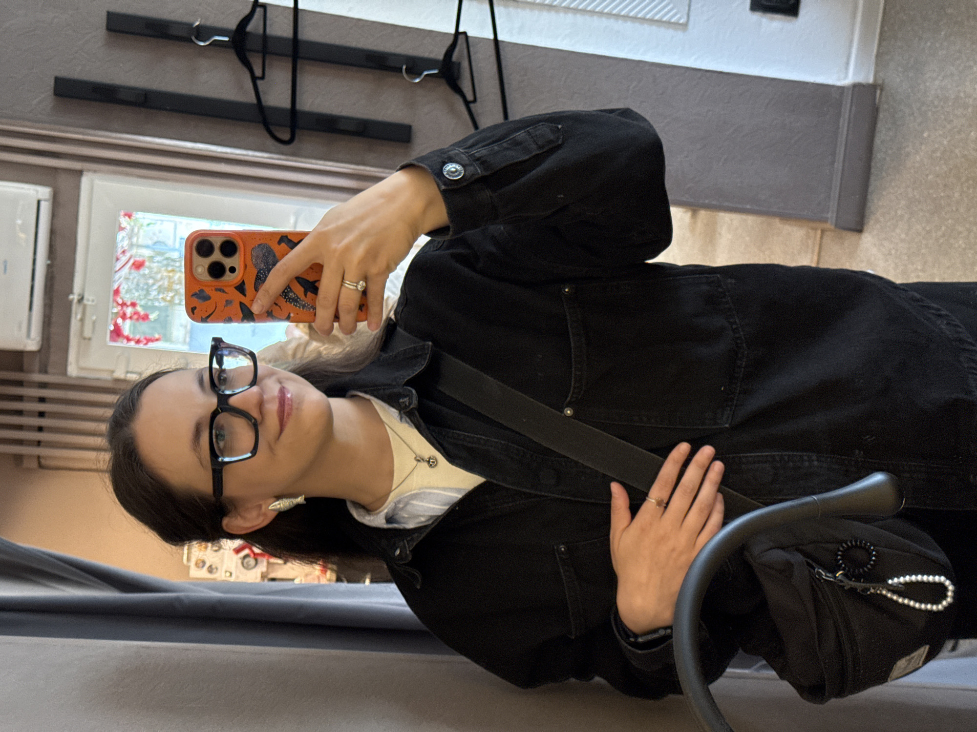 A young woman with dark hair and glasses is taking a mirror selfie in what appears to be a fitting room. She is wearing a black denim jacket over a light-colored shirt, with a necklace and earrings. She holds an orange-cased smartphone in her right hand, and her left hand rests on her waist. A black crossbody bag with a pearl keychain is visible. The background shows a grey curtain, a mirror, clothing racks with hangers, and part of a doorway with floral decorations. The environment suggests a retail setting, possibly a clothing store. There are no discernible features that would pinpoint this location to the city of Chaikovsky, Russia.