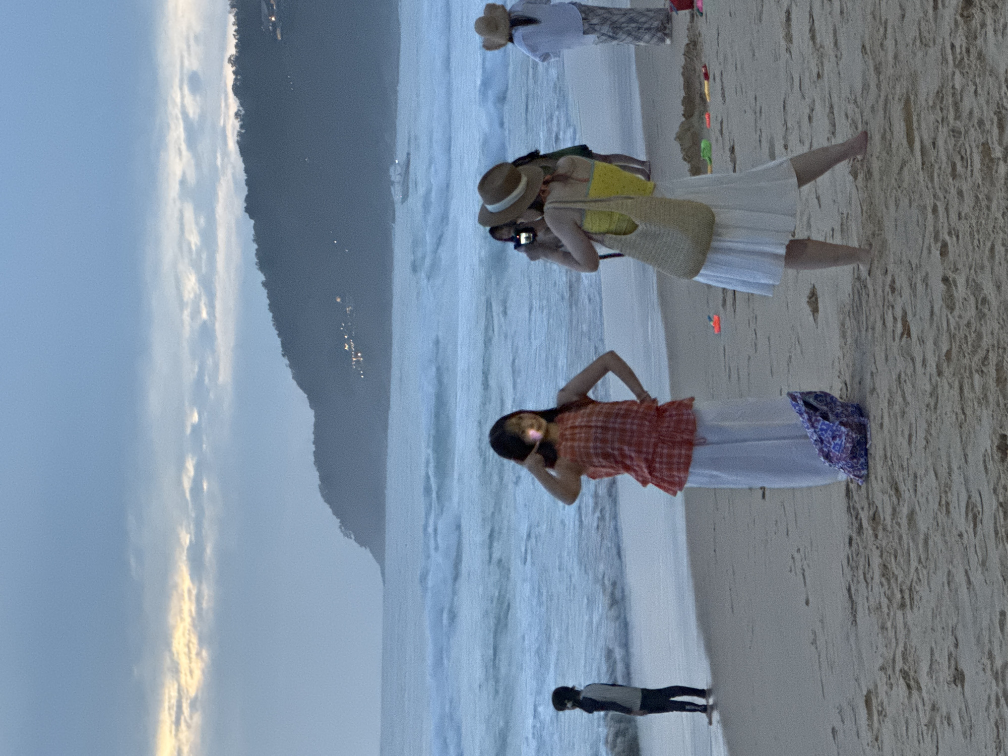 On a sandy beach in Karon, Thailand, several people are engaged in various activities as dusk settles. In the foreground, a woman in a yellow top and a white skirt stands with a straw bag, facing a person taking her photo with a camera. To her left, another woman in an orange and white plaid top and white pants poses with her hand near her face, illuminated by a light source. Further back and to the left, a person in dark clothing stands near the water's edge. In the middle ground, the ocean churns with waves, and a boat is visible in the distance. A hilly, tree-covered landscape with scattered lights forms the background, under a cloudy sky. Scattered on the sand are colorful toy beach accessories.