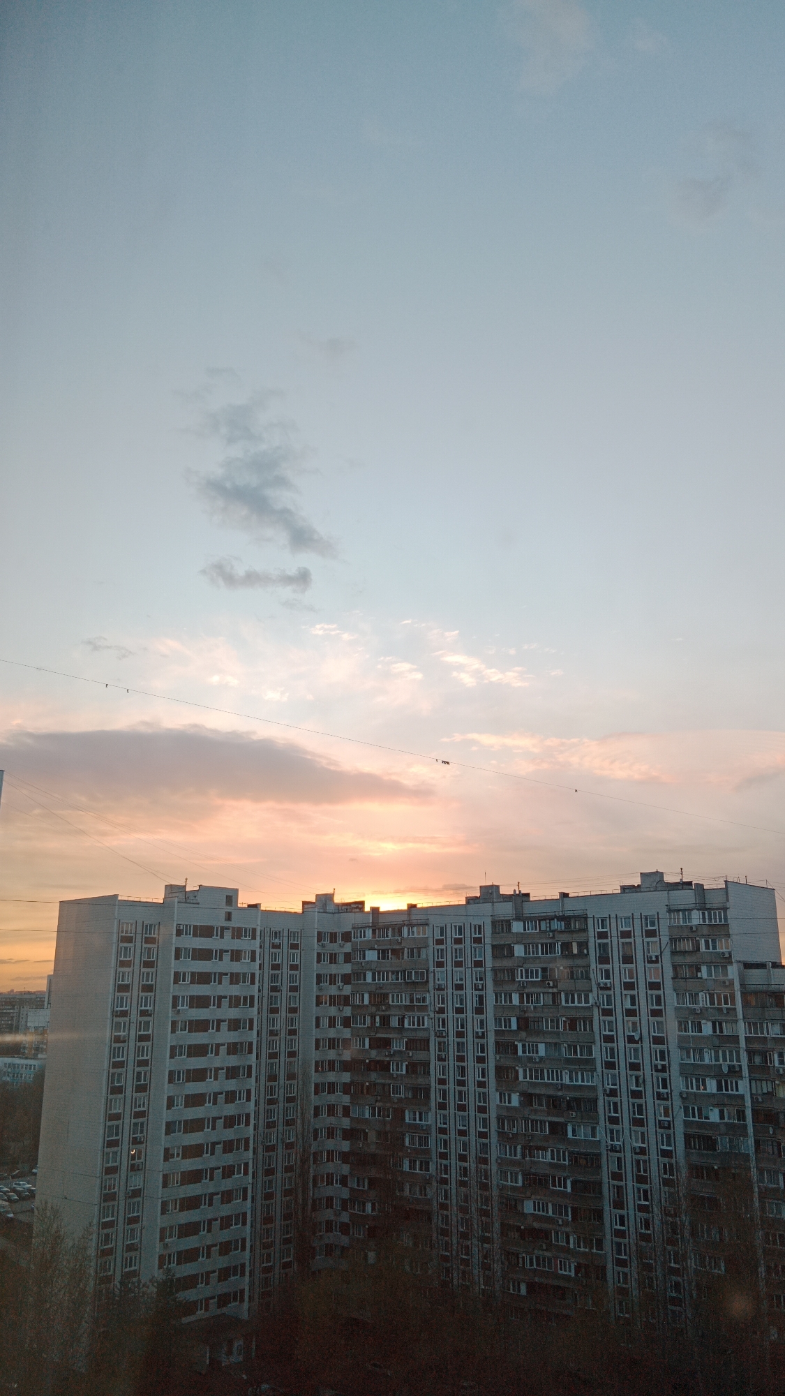 The image depicts a large apartment building in Moscow, Russia, likely from a high vantage point. The building is multi-storied with a repetitive pattern of windows and balconies, typical of Soviet-era architecture. A vibrant sunset fills the sky above the building, casting a warm, orange glow on the clouds. A few faint telephone or electrical wires stretch across the sky. The ground level is obscured by trees and vegetation. There is no evidence of people or specific actions in the image. A parking area with several cars is visible on the left side of the building. The environment suggests an urban setting in Moscow.