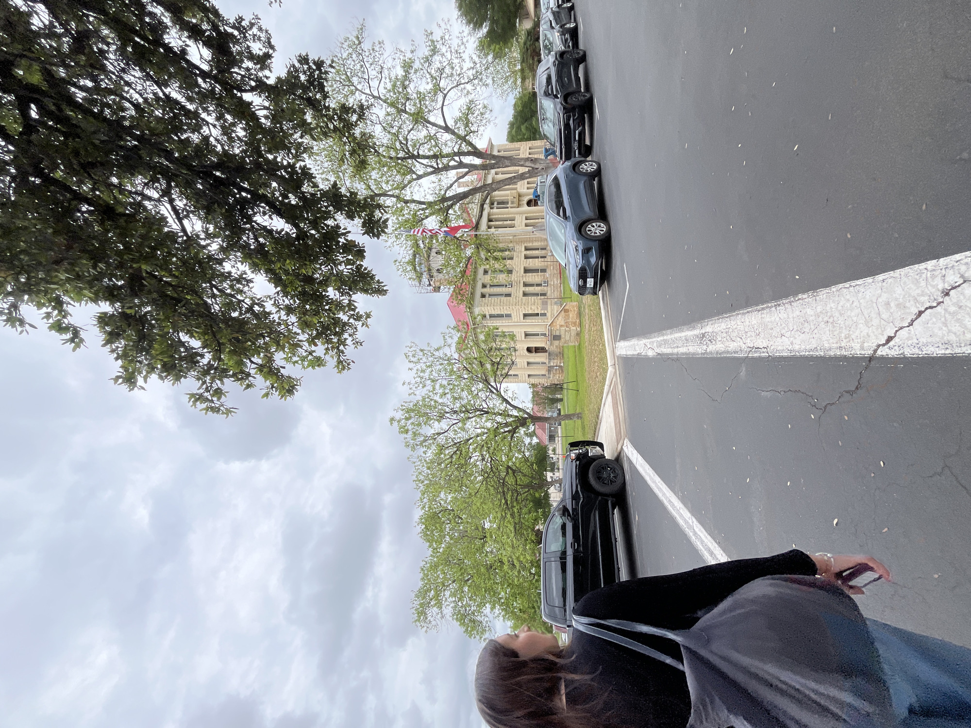 A ground-level, exterior shot, rotated 90 degrees counter-clockwise, shows a woman standing near a row of parked cars in Johnson City, United States of America.
The woman, partially visible in the lower right corner, has dark hair and is wearing a black shirt with a backpack. She is holding a mobile phone in her right hand.
A row of parked cars lines the right side of the shot, including a dark gray sedan, a dark SUV, and a black pickup truck. A weathered white parking line extends from the bottom towards the center of the frame.
In the background, a light-colored stone building, possibly a historical courthouse, stands amidst green trees. A flagpole displaying the American flag is visible near the building.
The sky is overcast with gray clouds. A large tree with dense green foliage overhangs the left side of the image.