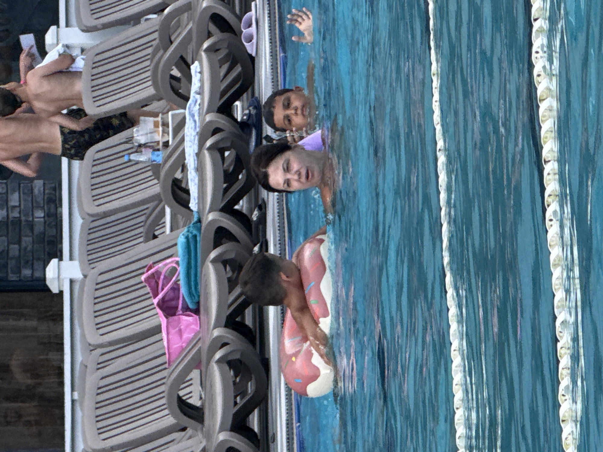 The image depicts a swimming pool area with several individuals in and around the water. In the foreground, two children and an adult woman are in the pool. One child, a young boy, is using a pink donut-shaped float. The woman is partially submerged, and another boy is visible behind her. In the background, multiple lounge chairs are set up, with some personal items such as a pink bag and folded towels on them. Two shirtless young men are standing behind the lounge chairs, appearing to interact with someone or something outside the frame. The environment suggests a warm-weather location, likely a resort or public pool facility. The location specified, Массандровский поселковый совет, Ukraine, implies a Crimean setting, which would align with the pool and lounge chair setup often found in resort areas.