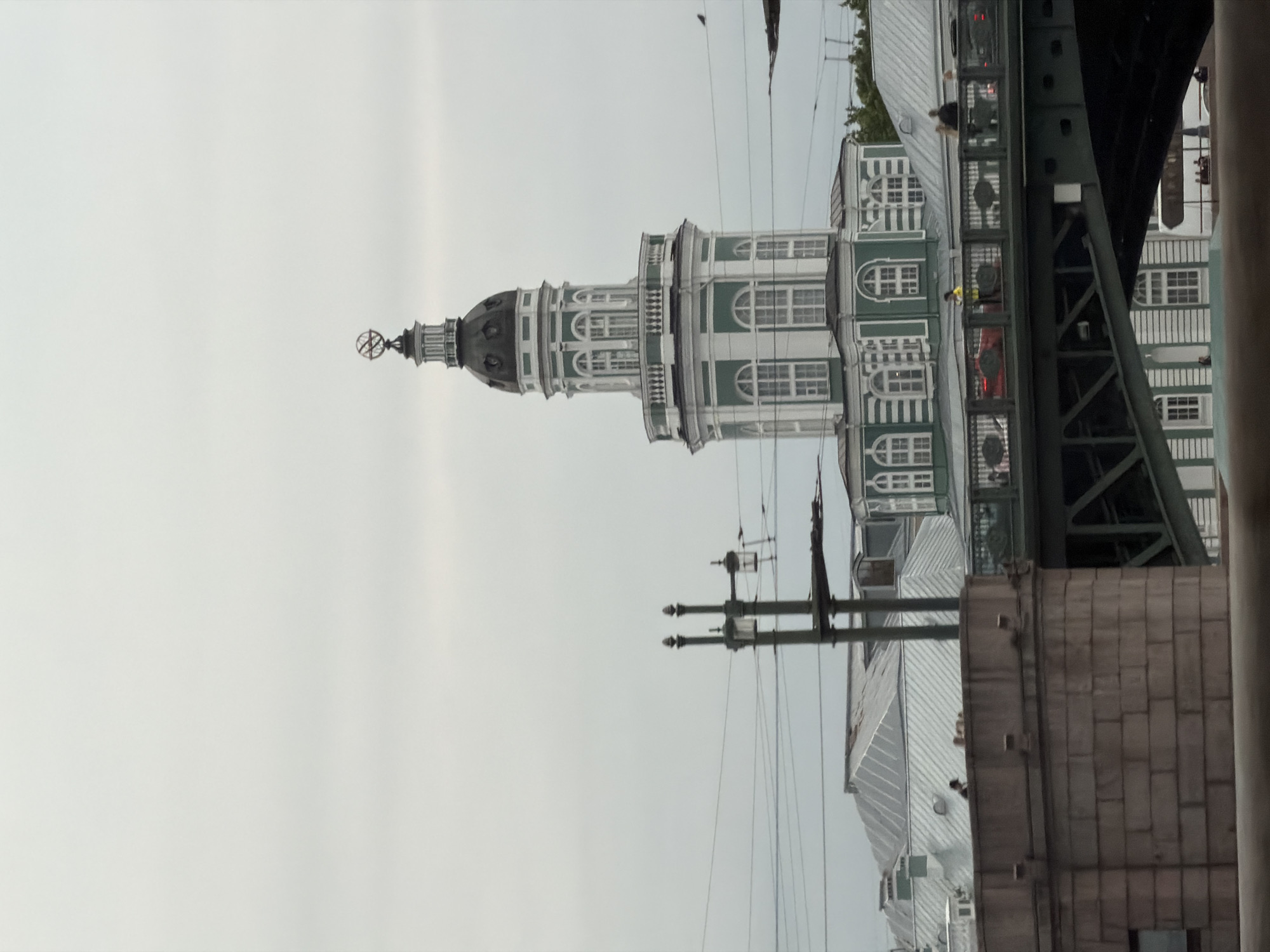 This is a medium shot, eye-level photograph of the Kunstkamera museum in Saint Petersburg, Russia, on an overcast day. The main subject is the museum's iconic tower, painted white and green, topped with a globe-shaped weather vane. In the foreground, a bridge crosses the frame, with people visible walking on it. The background features a cloudy sky and other buildings with distinctive architecture. The lighting is diffused and the overall color palette is muted due to the overcast weather.