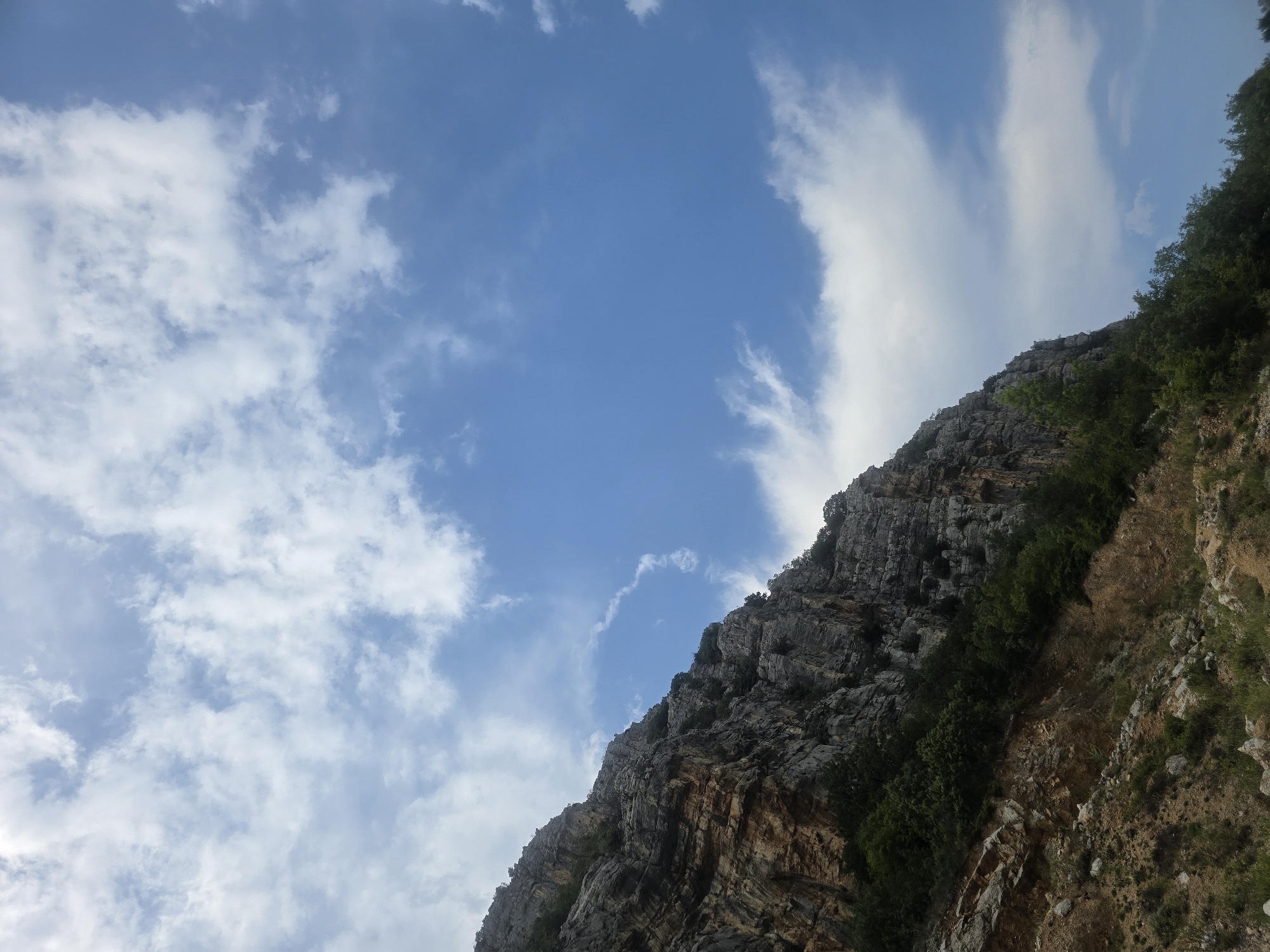 The image shows a steep, rocky mountainside under a bright blue sky with scattered white clouds. The mountainside is covered in patches of green vegetation, including trees and shrubs. The rocks are a mix of grey and brown, with visible layers and textures. The sky is a clear blue, with dynamic cloud formations suggesting a partly cloudy day. The angle of the shot is from below, looking up at the imposing mountain face. The foreground shows the lower part of the slope, with loose rocks and sparse vegetation. This scene is likely located in the mountainous region surrounding Podgorica, Montenegro, which is known for its rugged terrain and natural beauty.