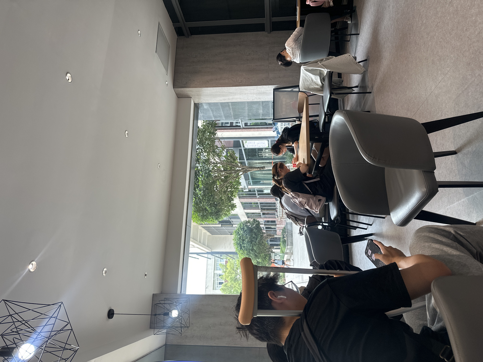A well-lit interior, likely a cafe or library, featuring several individuals. Seated patrons are engrossed in their activities; one person is on their phone, a crutch is visible resting against a nearby chair. Through a large window, a tree-lined street outside with visible buildings can be seen. The location is possibly in the Chongqing High-tech Zone, China.