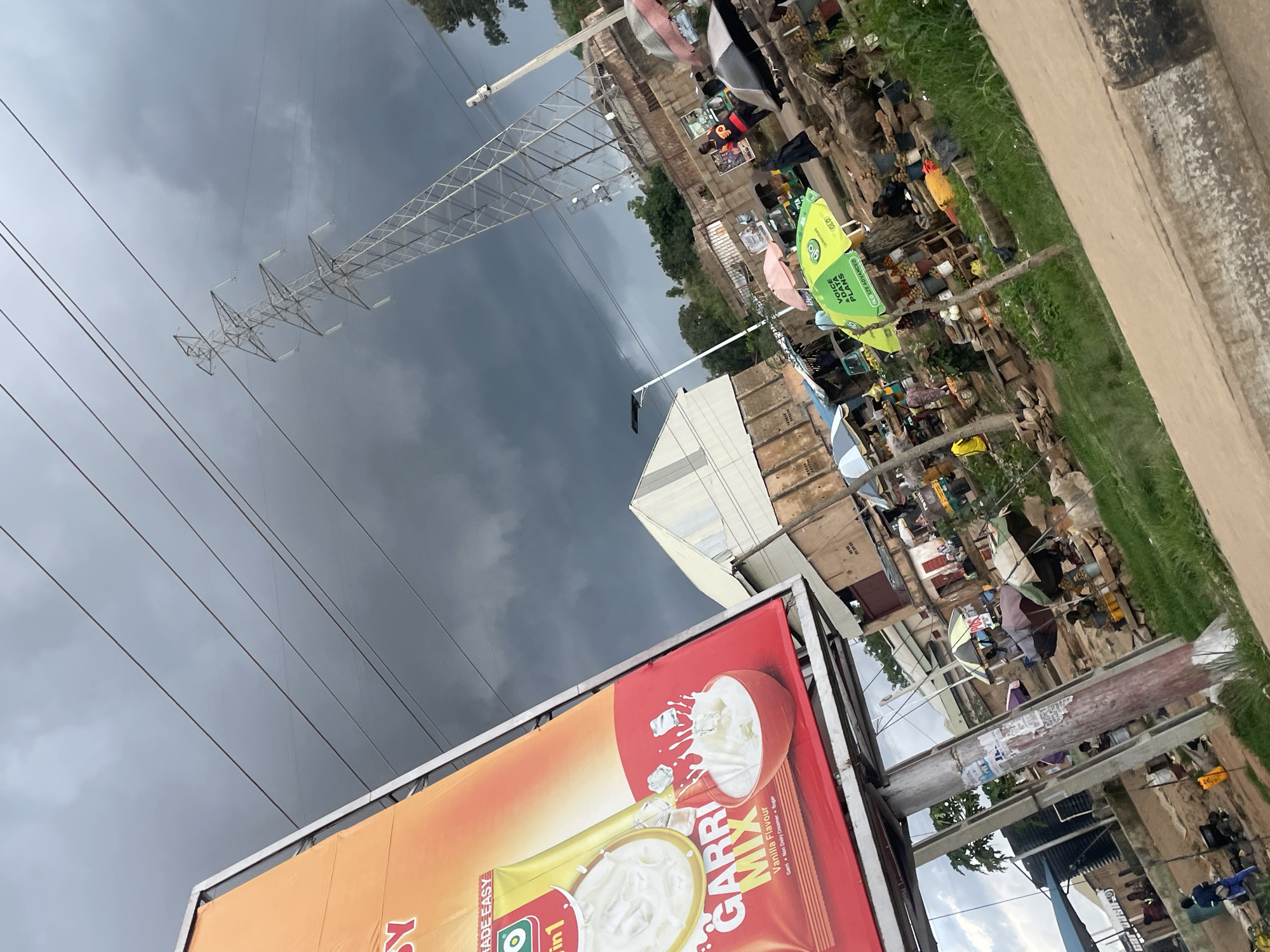 This image captures a street scene in Gura Top, Nigeria, under a dramatic, dark, cloudy sky. In the foreground, a large billboard advertises "GARRI MIX," a food product, with an image of milk being poured into a bowl. The billboard is supported by metal poles, one of which is covered in peeling posters. To the right of the billboard, a tall, lattice metal electricity pylon dominates the skyline, with thick power lines stretching across the frame.
The midground reveals a bustling marketplace. Various stalls are set up, some covered by colorful umbrellas, including a prominent green umbrella with "GLO VOICE & DATA PLANS" branding. People are visible tending to their stalls, shopping, or moving through the market. Goods are displayed on tables and in baskets, suggesting produce and other merchandise. Buildings with simple architecture line the background, some appearing weathered. A few vehicles, including a tricycle-like vehicle, are present in the market area. The ground is a mix of dirt paths and grassy areas. The overall impression is of a vibrant, active community scene, set against a backdrop of ominous weather and essential infrastructure.