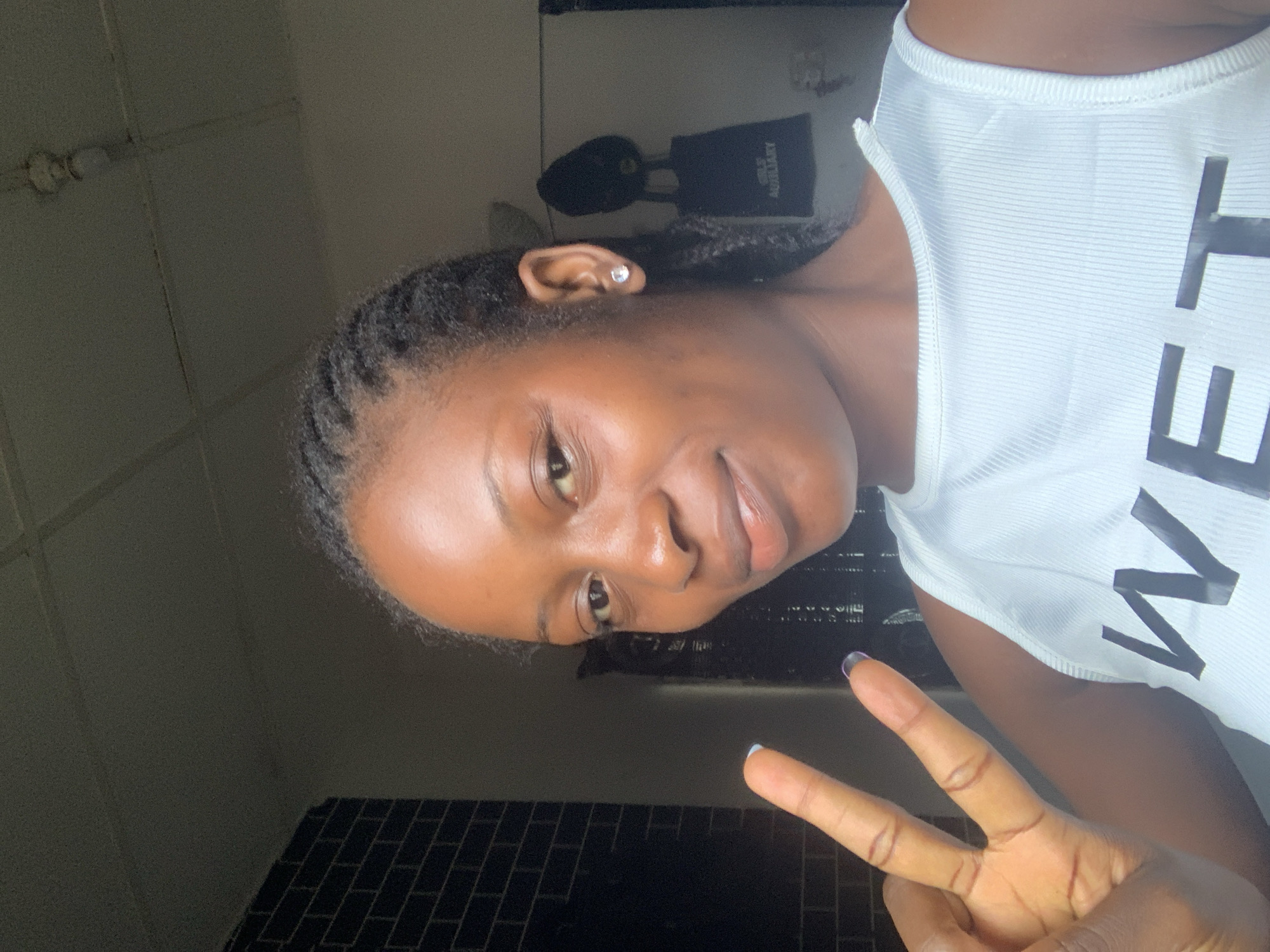 A dark-skinned woman with braided hair is positioned in the center-right of the frame, oriented sideways. She is wearing a white tank top with black letters and is making a peace sign with her right hand. The background appears to be a dimly lit interior space with tiled walls. A pair of headphones is visible on a shelf behind her. The location of the photo is in Lokoja, Nigeria.