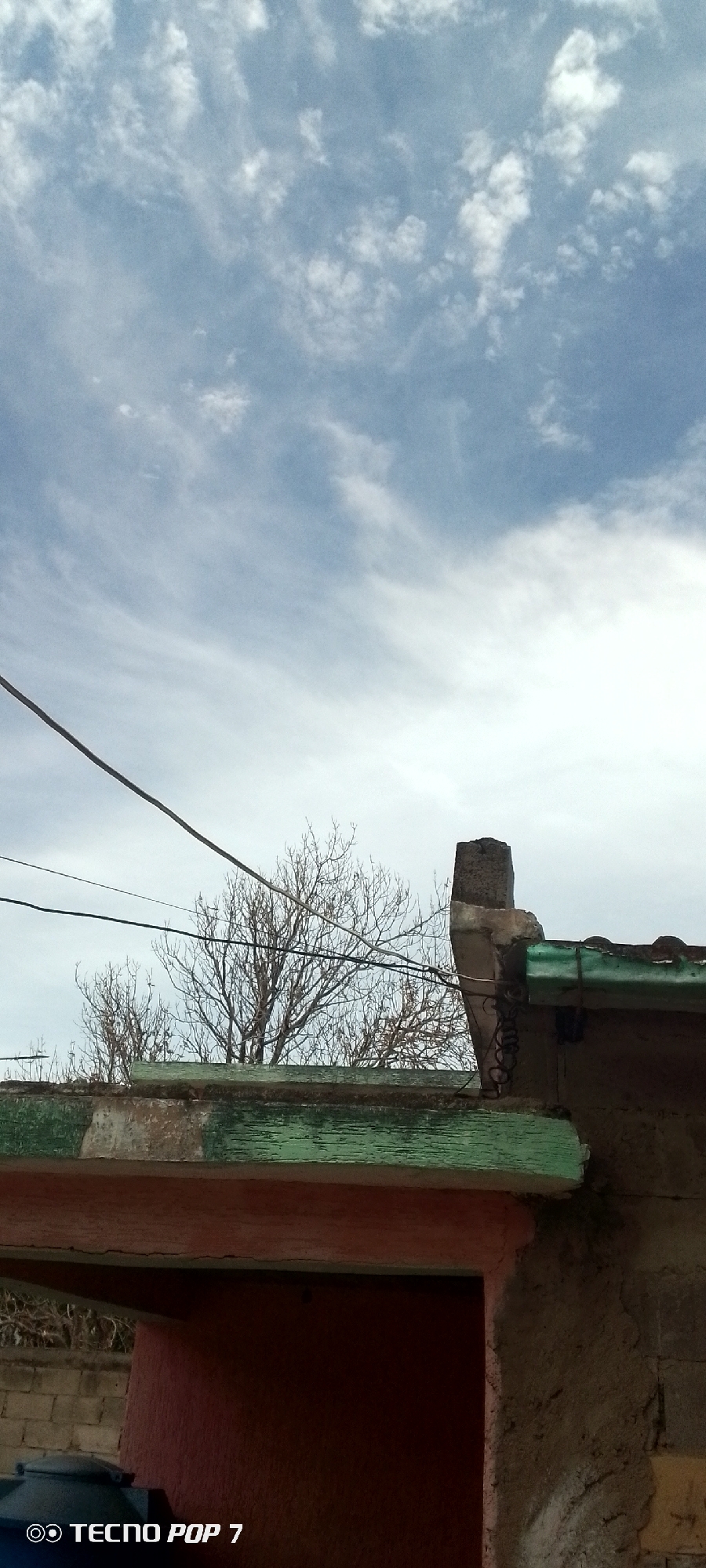 The image captures a daytime scene under a partly cloudy sky. A bare tree is visible behind a roof or a building structure with green trim. An electrical wire crosses the upper left of the frame. The structure below includes what appears to be a red-painted section above a water tank and a textured concrete wall. "Tecno Pop 7" is faintly visible in the bottom left corner. The location is unspecified, but the prompt suggests it is in the city of Altagracia, Venezuela. There are no people depicted in the scene.