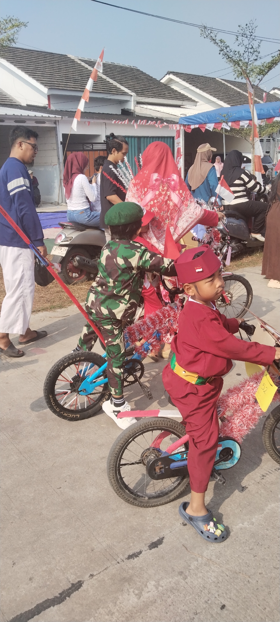 The image depicts a street scene in what appears to be a residential area. The context suggests a local event or celebration, possibly in Taban, Indonesia, given the presence of Indonesian flags and festive decorations. In the foreground, two young boys are positioned on bicycles. The boy on the left is wearing a camouflage military uniform and a green beret. The boy on the right is dressed in a red uniform with a matching red cap. Both bicycles are adorned with red and white streamers, reminiscent of national colors.

Behind the children, a group of adults and other children are visible. One woman is seated on a motorcycle, wearing a red hijab. Another adult is standing nearby, pulling the red and white pole attached to the boy's bicycle on the left. Several other individuals are standing and observing, some partially obscured by the main subjects. The background shows houses with tiled roofs, characteristic of many urban and suburban environments. The ground is paved, suggesting a road or street. The lighting indicates it is daytime, with clear skies. The overall atmosphere appears to be one of a community gathering, likely related to a national holiday or local parade.