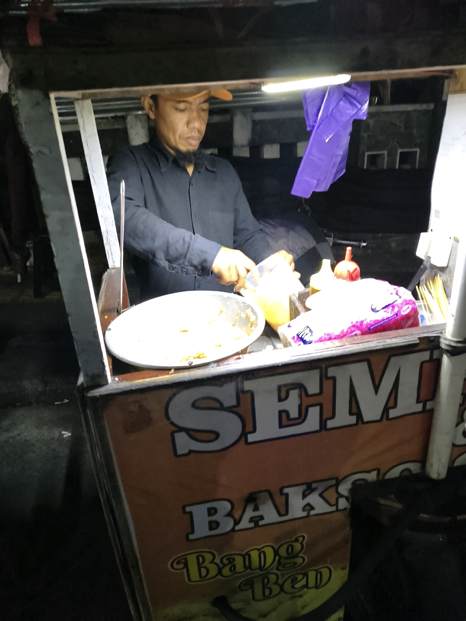 A man wearing a black shirt and an orange cap is preparing food at a street cart at night. The cart has an orange banner with text that reads "SEM" and "BAKSO". There are various food items and condiments on the cart, including a bowl of food, a plastic bag of what appears to be snacks, and bottles of sauce. A fluorescent light illuminates the scene. The background is dark, suggesting it is nighttime, and shows some buildings and structures. The overall environment appears to be an outdoor setting, likely a street in Bandungan, Indonesia, given the context of the image.