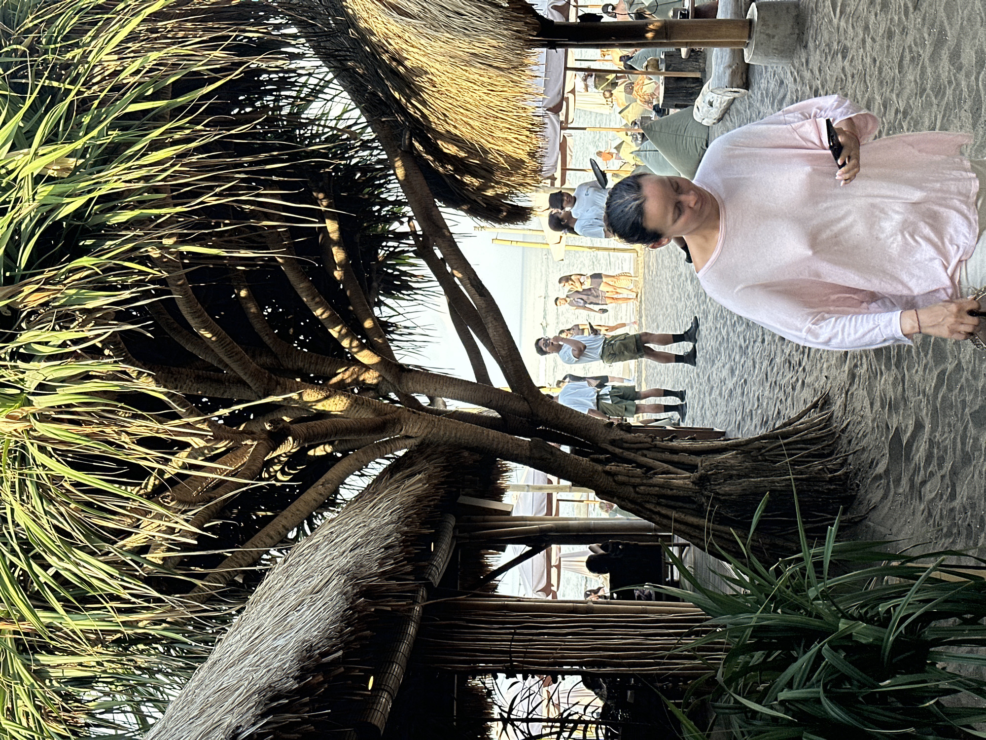 A woman in a light pink, translucent long-sleeved shirt and light-colored pants walks on a sandy beach in North Kuta, Indonesia, holding a smartphone in her right hand. She has dark hair pulled back and is looking down at her phone. Behind her, several people are visible on the beach, some wearing casual clothing. Two thatch-roofed structures with bamboo detailing are present, and a large tree with exposed roots stands between them. Beach umbrellas and seating areas are also visible in the background, suggesting a resort or dining establishment. The scene is illuminated by natural sunlight, casting shadows on the sand.