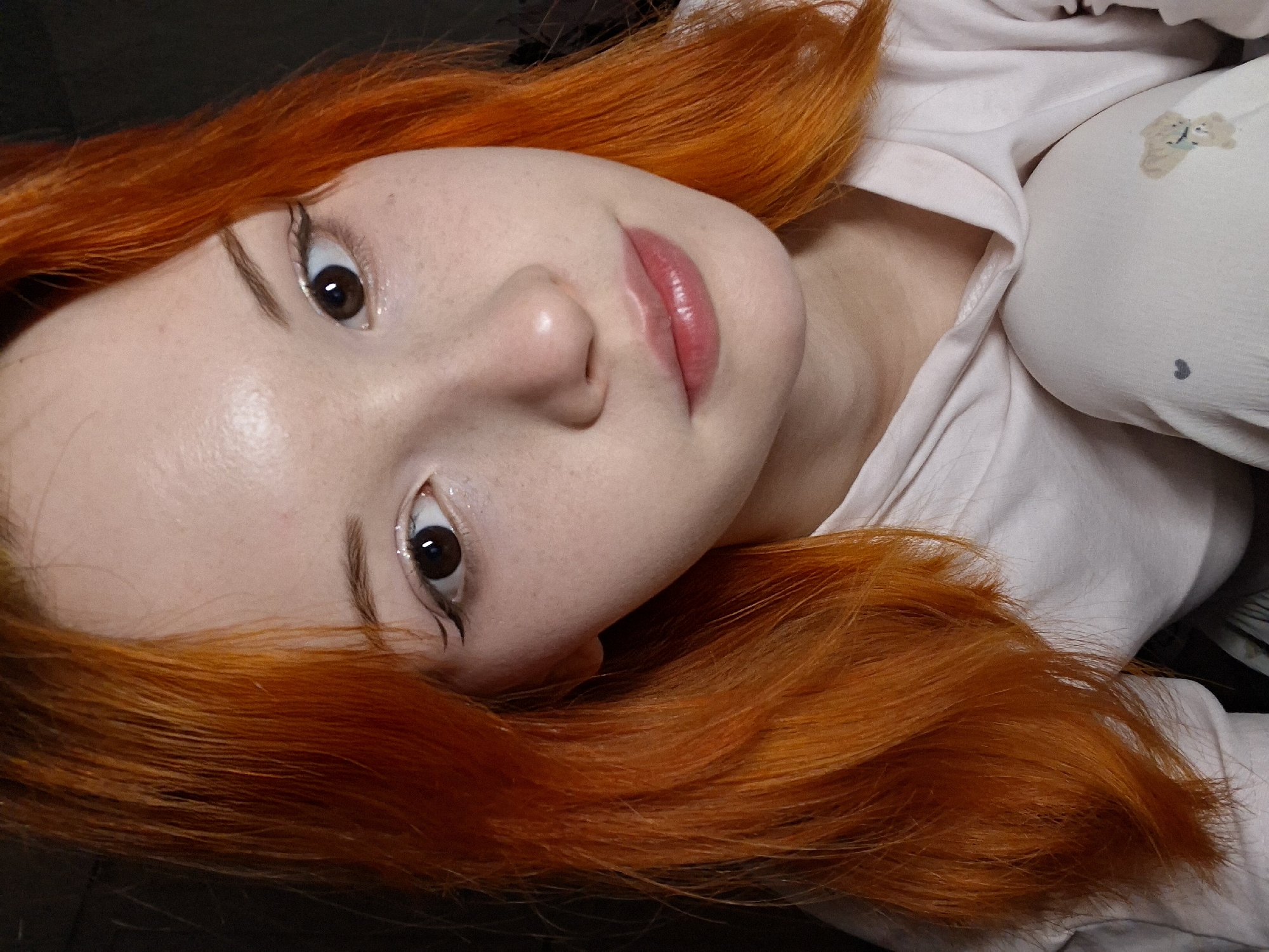 An overhead portrait features a young individual with striking, vibrant orange-red hair, fair skin, and dark brown eyes, lying down and looking towards the camera. The person's face occupies the left and central part of the frame, with their head tilted. Their hair is fanned out around their head against a dark background, likely a dark surface or bedding.
Notable facial features include wide-open eyes with a slight gleam, accented by winged black eyeliner. Freckles are visible across the forehead and cheeks. The skin appears smooth and slightly dewy. The lips are full and tinted with a glossy, light pink or peach color.
The person is wearing a light-colored (likely white or off-white) soft garment, possibly a hoodie or comfortable top, which is visible around their shoulders and upper chest. A small, faint, light-brown teddy bear illustration is present on the fabric near the right shoulder area, with a tiny dark speck below it. The setting appears to be indoors, suggesting a relaxed environment like a bed or couch. The overall mood is calm and direct. This image was captured in Chaikovsky, Russia. No visible text is present.