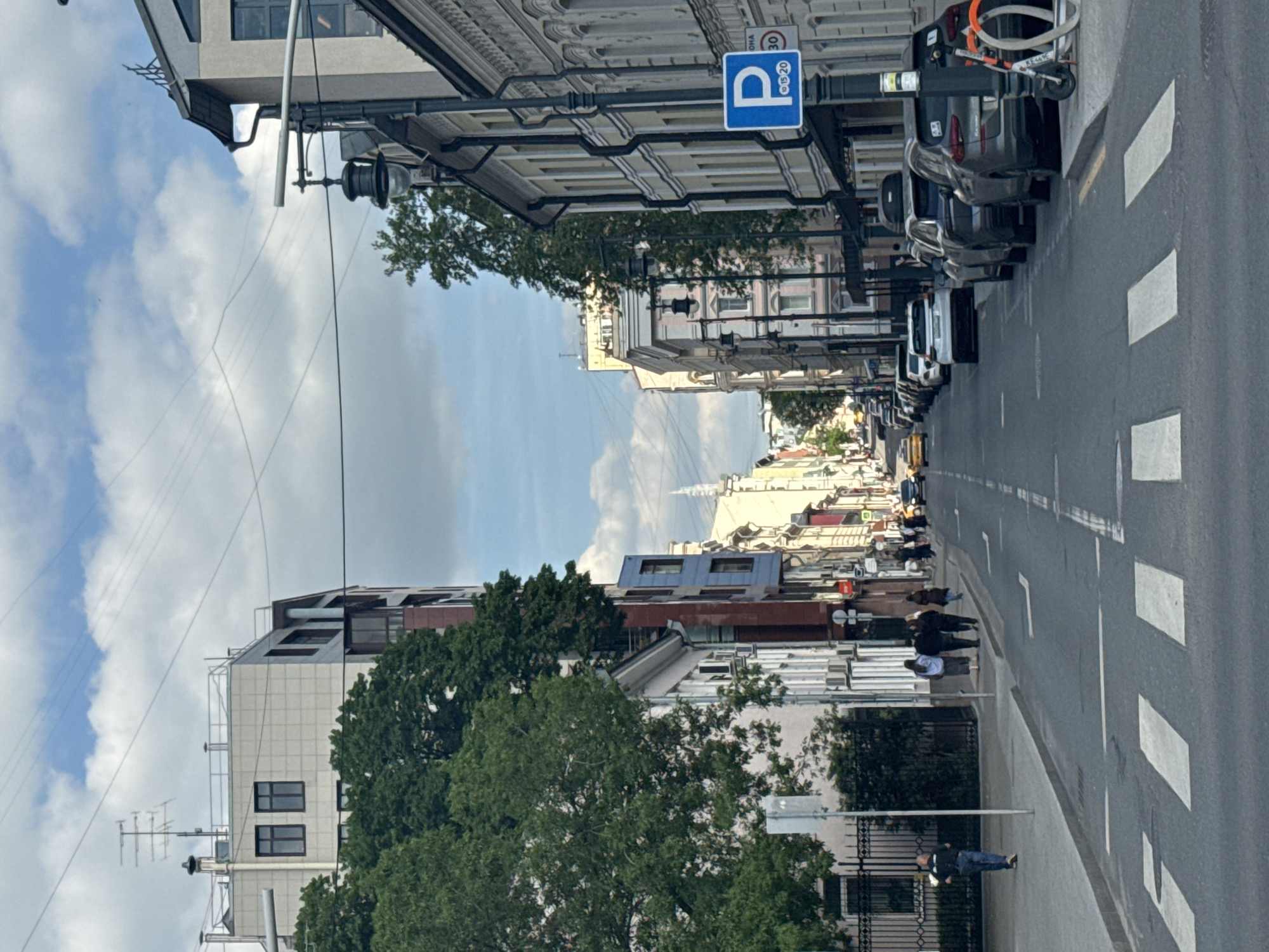 The image captures a street scene in Moscow, Russia. The view looks down a straight avenue lined with buildings of varying architectural styles, indicative of different historical periods. The sky is partly cloudy. The street itself has white pedestrian crossing markings and is lined with parked cars on the right side. People are walking along the sidewalk on both sides of the street. A blue parking sign is visible on the right side of the street. Trees are seen on the sides, adding greenery to the urban landscape. The general environment suggests a typical day in the city with pedestrian and vehicular activity.
