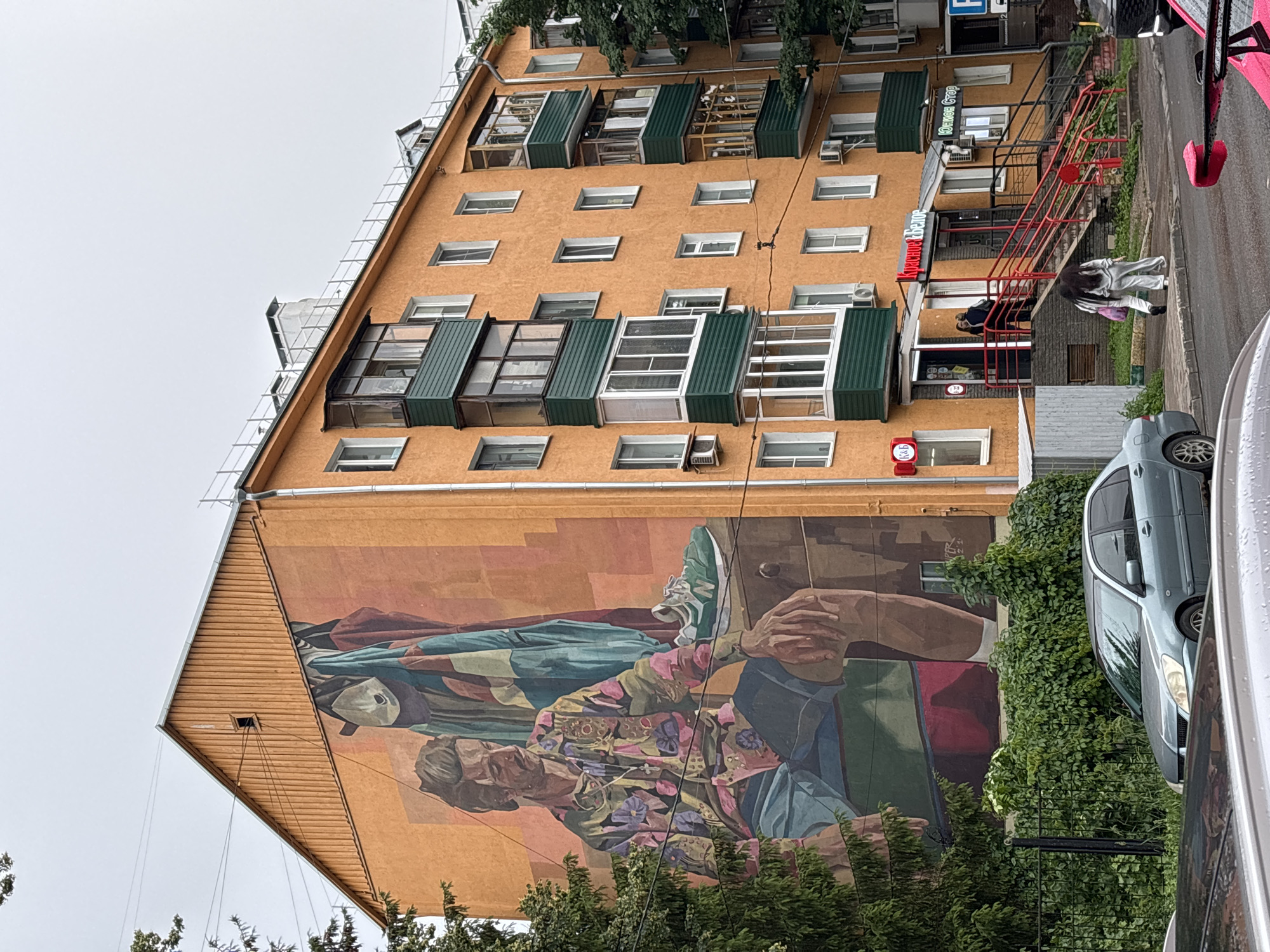 The image shows a mural on the side of an orange apartment building in Nizhny Novgorod, Russia. The mural depicts a man in a colorful, patterned shirt and shorts, sitting with his arm resting on a piece of furniture. A green sneaker and a jacket are placed on top of the furniture. The building has several balconies, some enclosed with glass and others open. Below the building, there is a sidewalk, a black metal fence, and lush green bushes. A light blue car is parked on the street in front of the bushes. Further down the street, a red and white awning with Russian text is visible above store entrances, and people are walking on the sidewalk and climbing red-railed stairs. The sky is overcast.