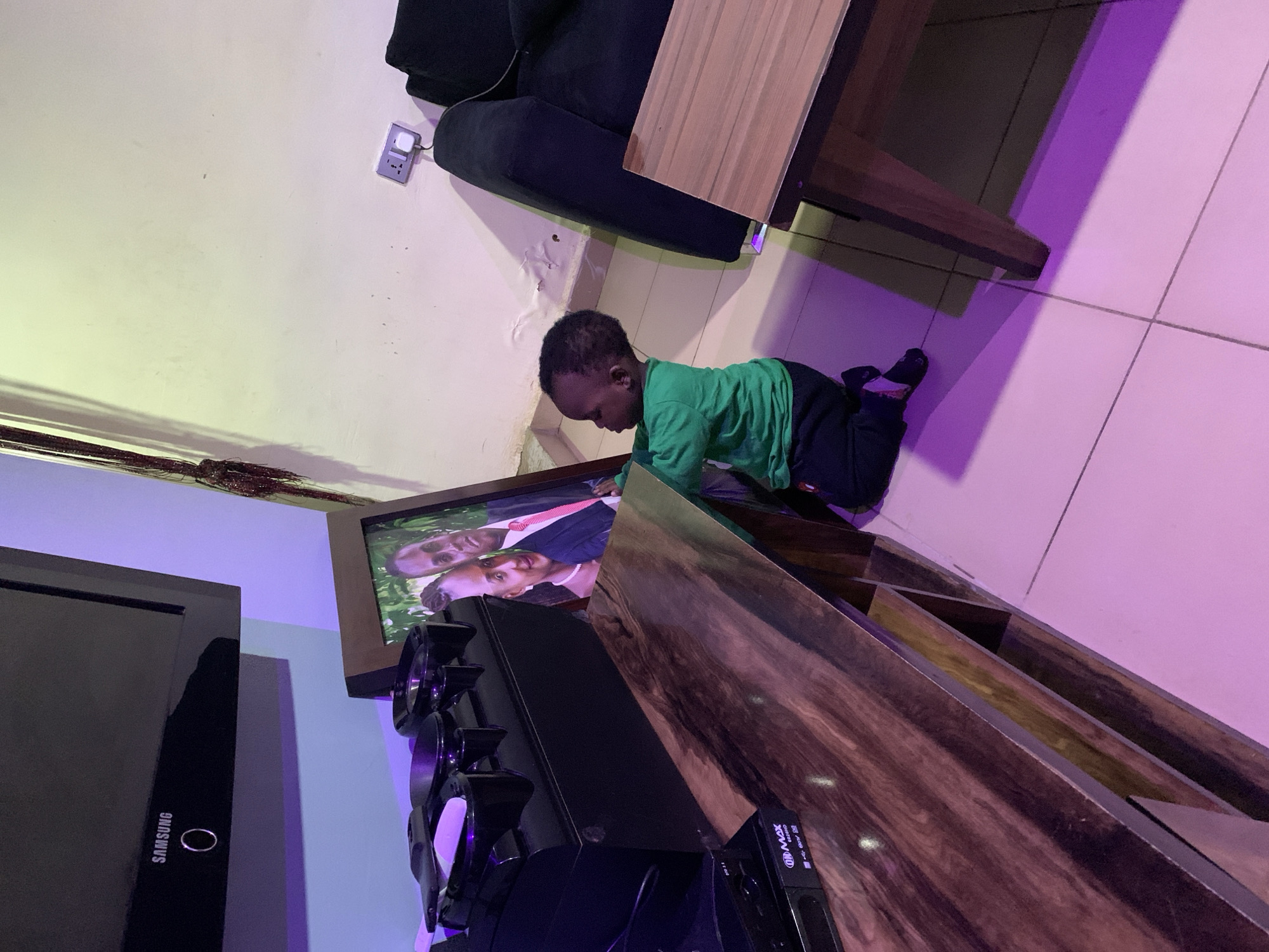 A young Black child wearing a green long-sleeved shirt and dark pants kneels on a light pink tiled floor, reaching towards a framed portrait of an adult couple. The portrait is leaning against a black entertainment center. The entertainment center holds various electronic devices, including a Samsung television mounted on the wall above it. The room is dimly lit, with purple and yellow lighting casting shadows. To the right of the child and portrait is a dark blue sofa and a wooden table with a lighter wood top. A wall socket with a plug is visible on the wall behind the sofa.