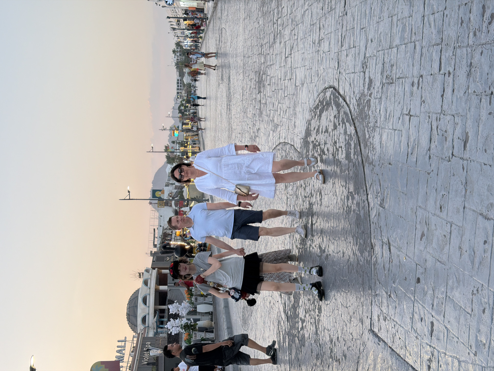 The image shows a street scene in Sharm El Sheikh, Egypt, during what appears to be sunset or early evening. The street is paved with large, textured gray stones. In the foreground, a woman in a white dress walks towards the viewer. Behind her, a man in a light blue polo shirt and dark shorts walks with a young person dressed in a gray t-shirt, black skirt, and patterned socks. The young person is holding hands with the man and carrying a stuffed toy and a small bag. To the left, another man in a black t-shirt and shorts is visible walking away from the viewer. The background features buildings with signs such as "MALL" and "Violet Spa," along with streetlights and palm trees, indicative of a commercial area. Several other people are scattered further down the street, suggesting a public thoroughfare. The sky is a soft orange and yellow hue, typical of a sunset.