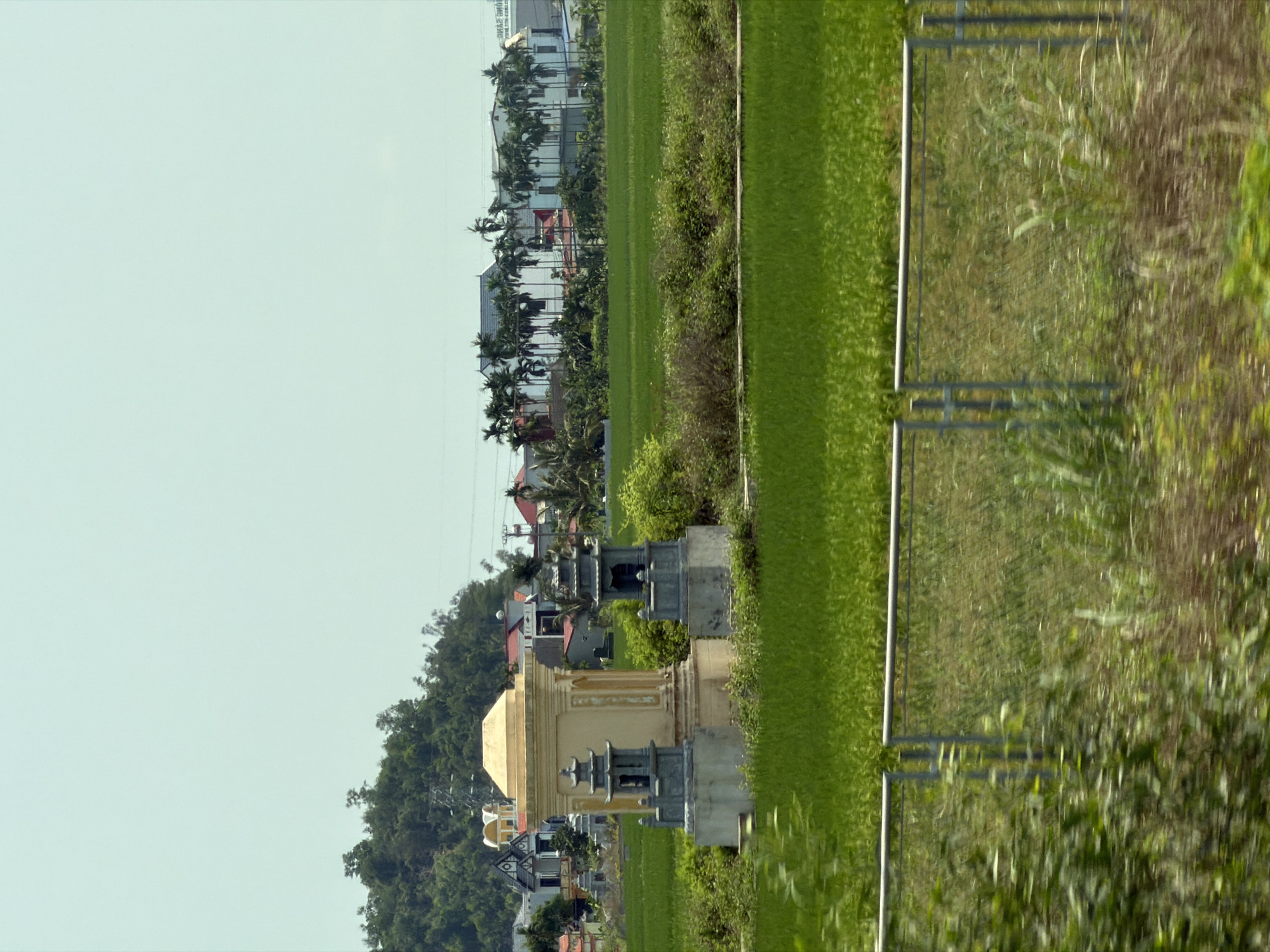 The image shows a landscape in what is likely a rural or suburban area of Hải Phòng, Vietnam. There is a field of green vegetation, potentially rice paddies, stretching across the middle ground. A row of houses and buildings, some with red roofs and palm trees, lines the horizon in the background. In the foreground, there is a partially visible metal fence separating the field from an area of taller grass and brush. Additionally, there are several structures that appear to be part of a cemetery. These structures are beige with grey or silver ornamentation. Some of these are decorated with traditional elements. The sky is pale and clear, suggesting daylight.