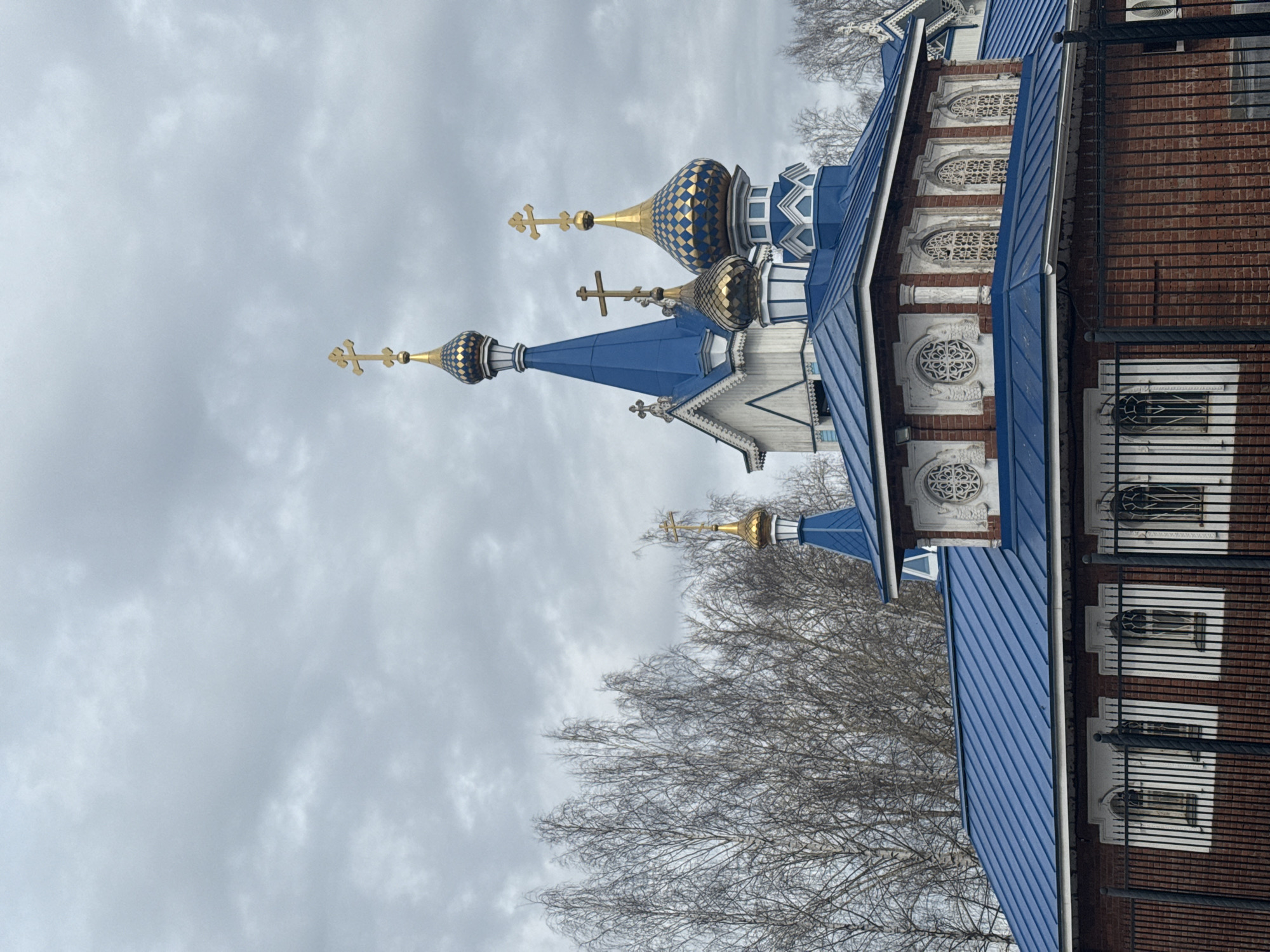 The image shows the exterior of the St. Michael's Cathedral in Izhevsk, Russia. The cathedral has a blue roof and several domes, some with gold and blue checkerboard patterns. Golden crosses top each dome. The building is constructed of red brick with decorative white stone elements, including arched windows and circular motifs. Bare trees are visible near the building. A black metal fence runs along the side of the structure. The sky is overcast.